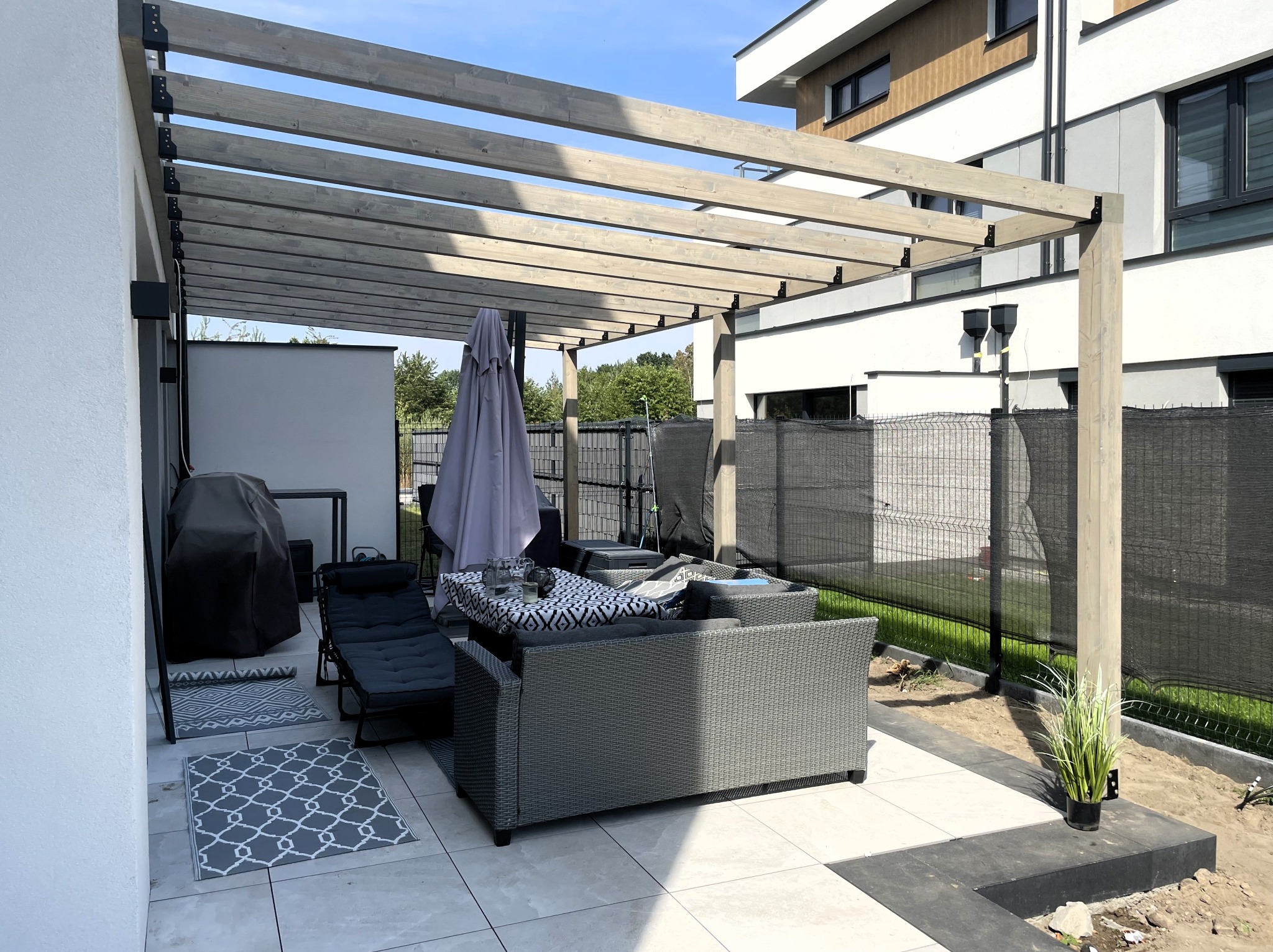 Nowoczesna pergola z jasnego drewna, z szarymi meblami ogrodowymi i roślinami w donicach. Widok na taras z jasnych płytek i fragment domu w tle.