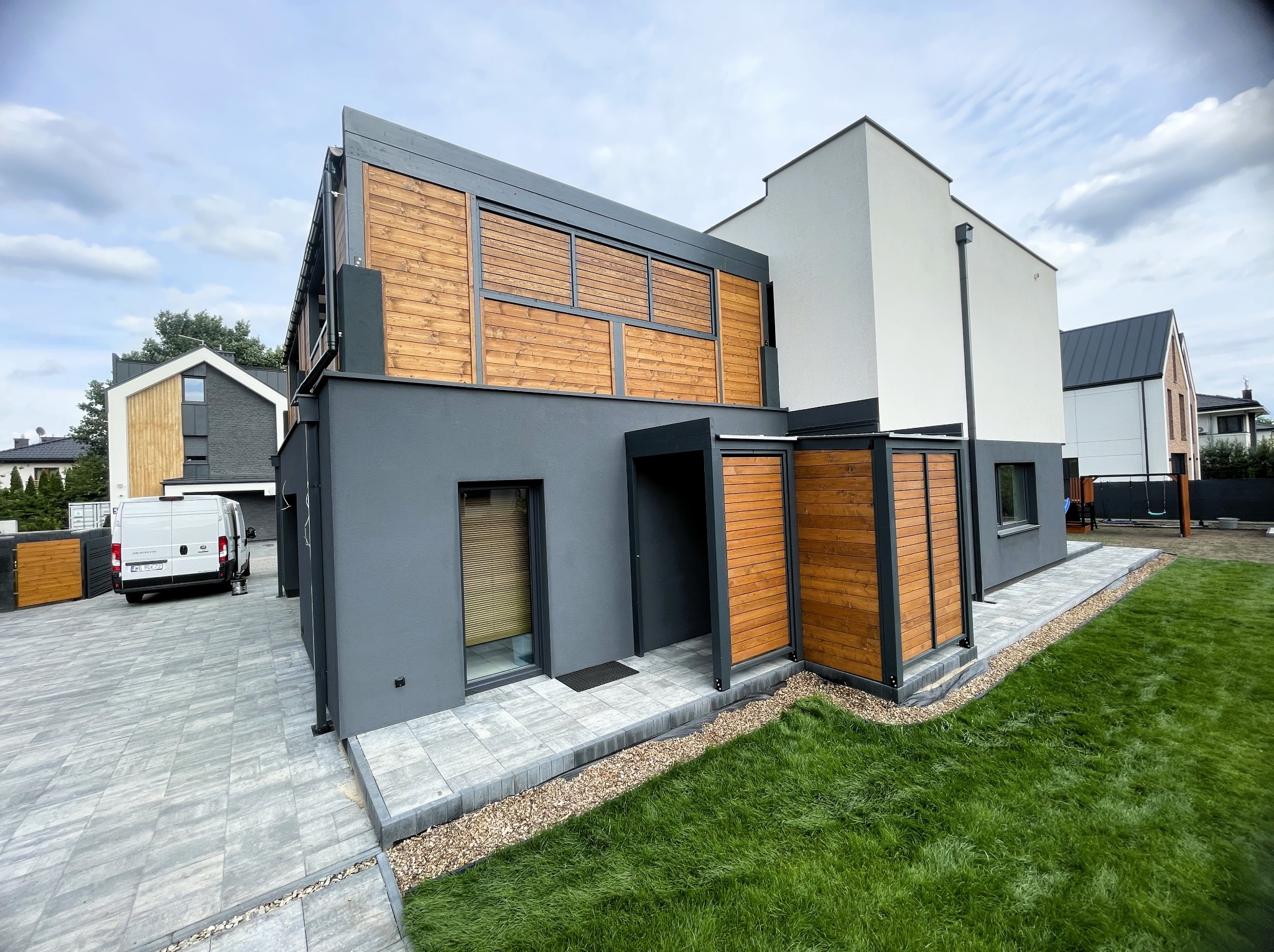 Nowoczesny dom z elementami drewnianymi: elewacja z szarego tynku i drewnianych paneli, kostka brukowa na podjeździe, zadbany trawnik. Minimalistyczny design.