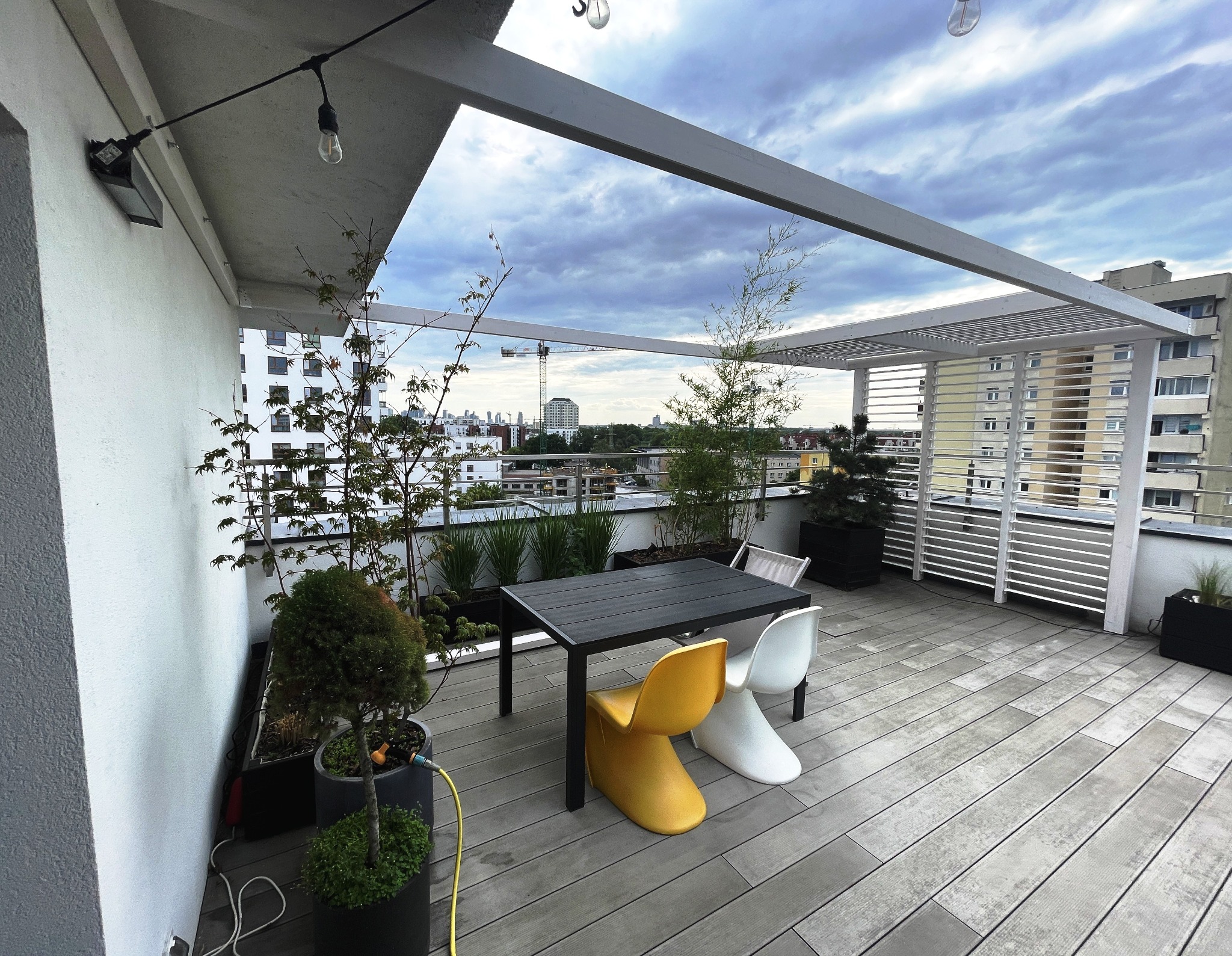 Nowoczesny taras na dachu z szarymi deskami, minimalistyczną pergolą i roślinami w donicach. W tle widok na miasto i pochmurne niebo. Żółte i białe krzesła przy stole.