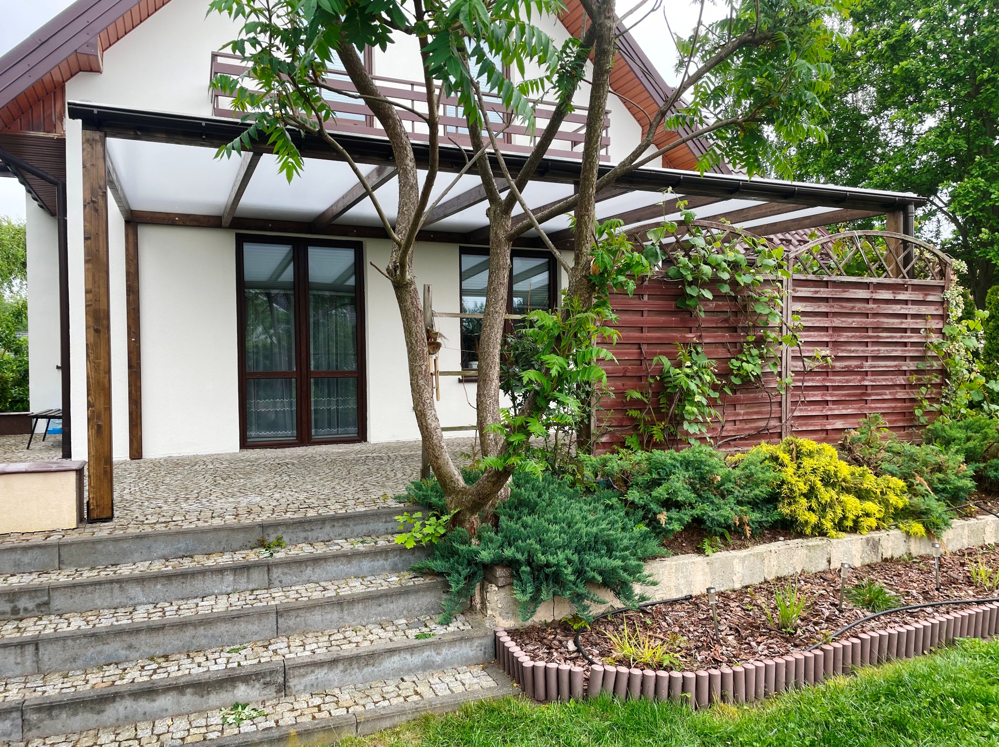 Dom z zadaszonym tarasem i drewnianym ogrodzeniem. Widoczne schody wejściowe z kostki brukowej i zadbany ogród z roślinami. Zadaszenie tarasu z drewnianą konstrukcją.