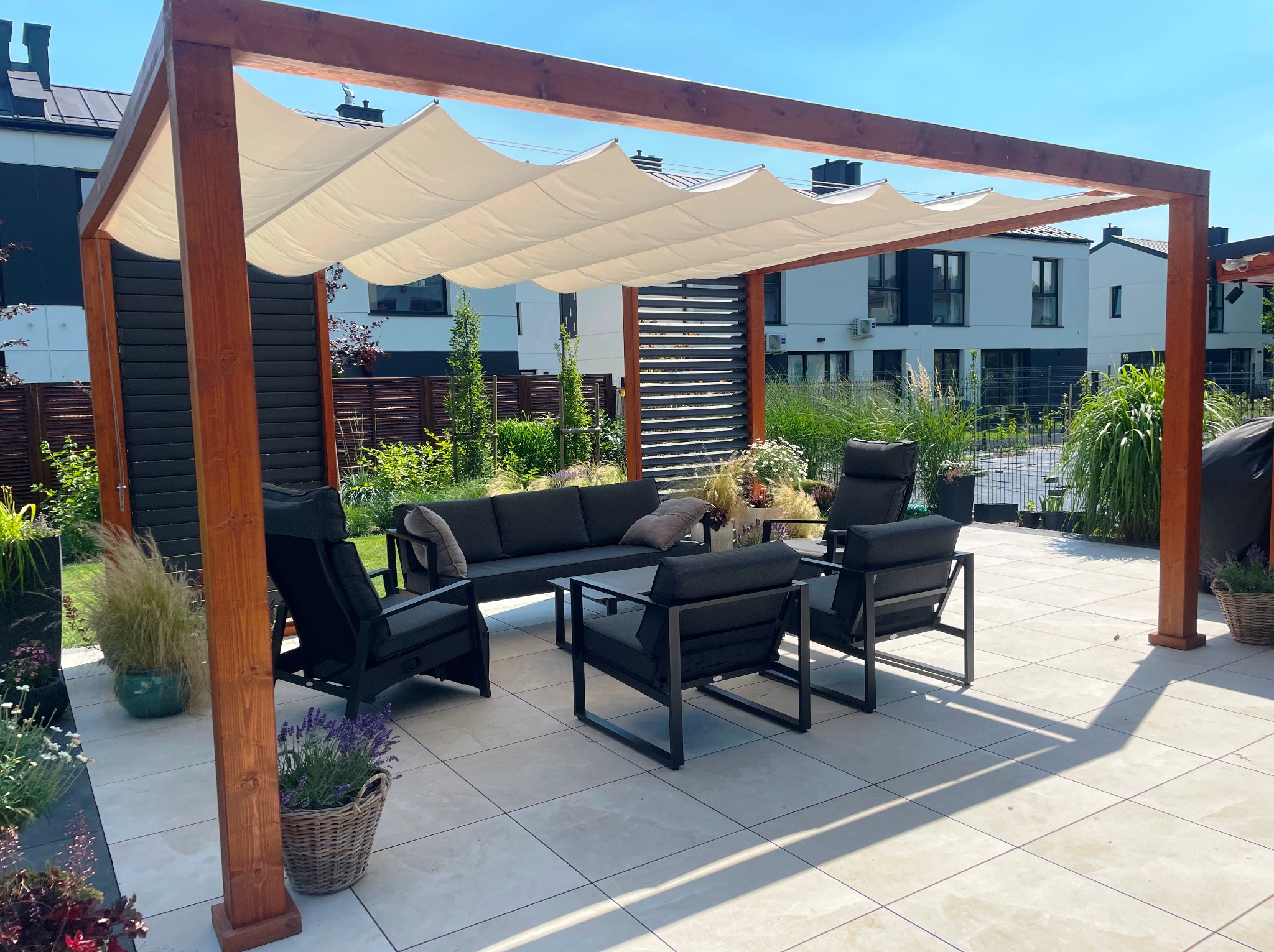 Nowoczesna pergola z drewnianą konstrukcją i kremowym, falistym zadaszeniem, otoczona meblami ogrodowymi na jasnym tarasie. W tle domy i zieleń.