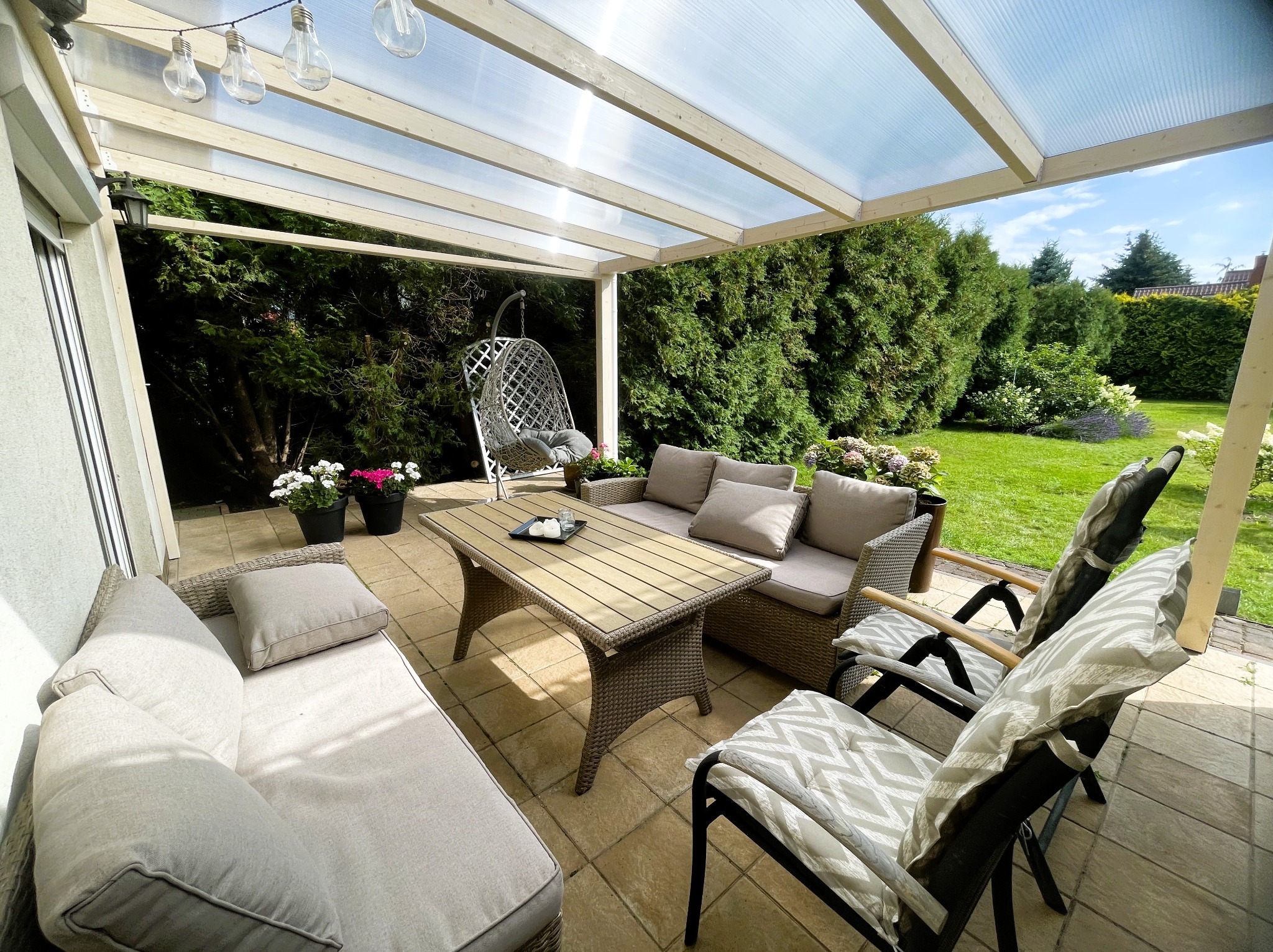 Zadaszony taras z meblami ogrodowymi w stylu rattan. Widoczna konstrukcja z przezroczystym dachem, wiszące lampki i huśtawka. W tle zadbany ogród z zielonymi krzewami.