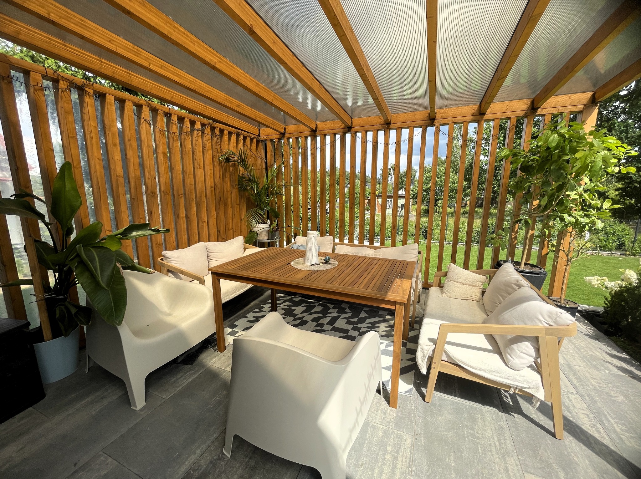 Zadaszony taras z drewnianą konstrukcją, wyposażony w meble ogrodowe: stół, krzesła i sofy z poduszkami. Na podłodze wzorzysta mata. Widok na ogród w tle.