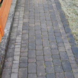 Mycie Ciśnieniowe Piotr Zieliński - Ciemna kostka brukowa, częściowo wyczyszczona, z widoczną różnicą w kolorze. Po prawej stronie trawnik, po lewej drewniany płot.