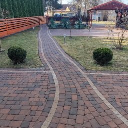 Mycie Ciśnieniowe Piotr Zieliński - Kaskadowa ścieżka z kostki brukowej w odcieniach brązu i beżu, otoczona zielenią trawnika i niskimi krzewami, prowadząca do drewnianej altany w słoneczny dzień.