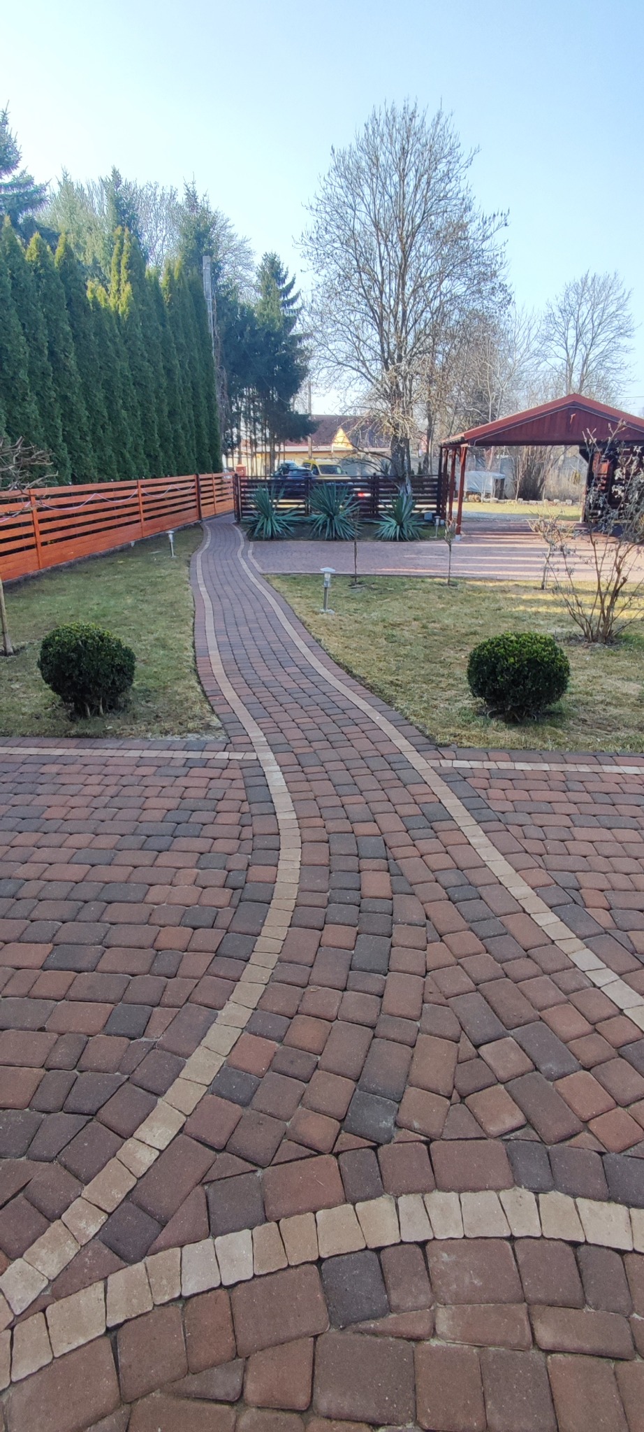 Kaskadowa ścieżka z kostki brukowej w odcieniach brązu i beżu, otoczona zielenią trawnika i niskimi krzewami, prowadząca do drewnianej altany w słoneczny dzień.