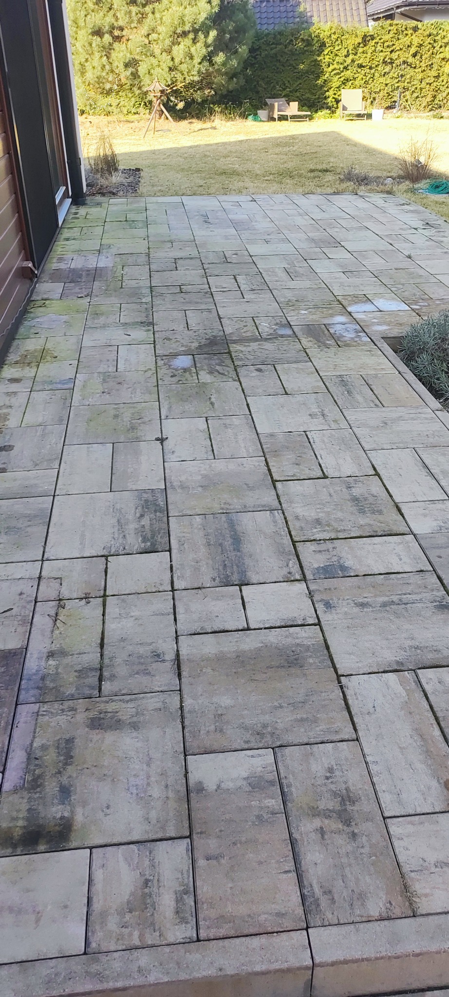 Brudna kostka brukowa na tarasie przed domem, widoczne plamy i zabrudzenia. Tło stanowi trawnik i elementy architektury ogrodowej.