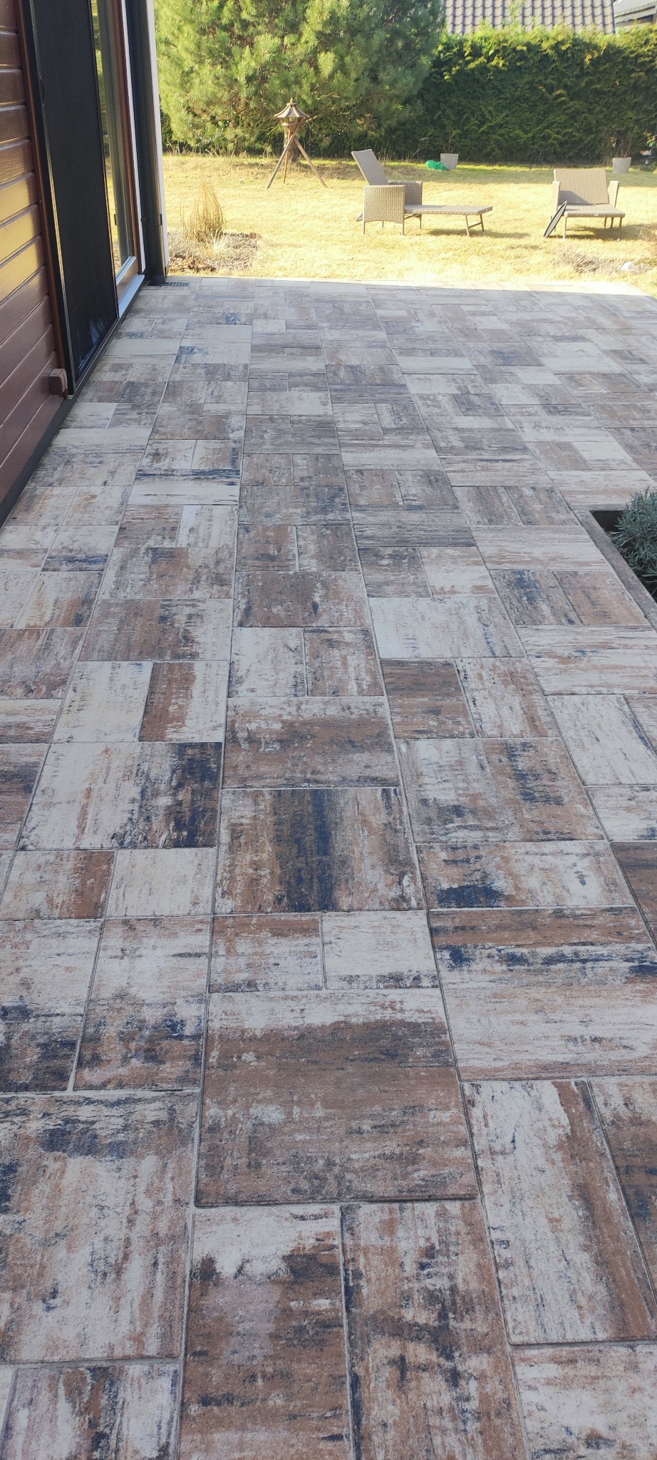 Ułożona kostka brukowa w odcieniach brązu, bieli i szarości na tarasie, z widokiem na trawnik i meble ogrodowe w tle. Widoczne detale faktury kostki.