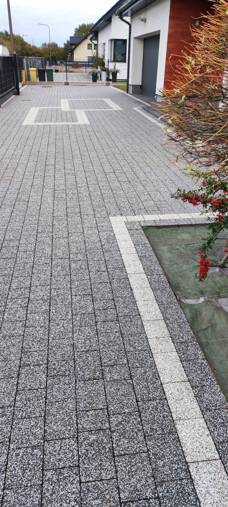 Szara kostka brukowa z białymi wstawkami na podjeździe, widok z góry, z elementami zieleni i fragmentem budynku w tle. Czysta powierzchnia, regularny wzór.