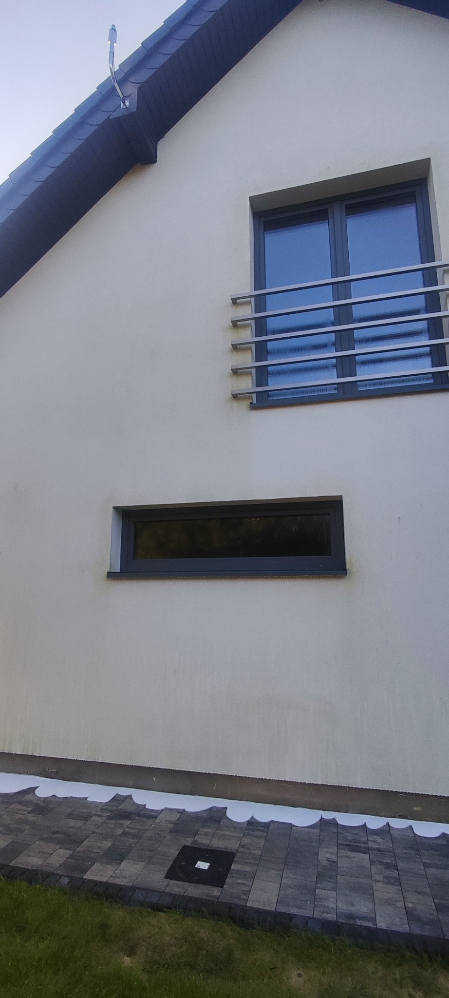 Jasna elewacja domu z dwoma oknami w szarych ramach, z czego jedno z metalową barierką. U dołu widoczna kostka brukowa i trawa. Widoczne zabrudzenia na elewacji.