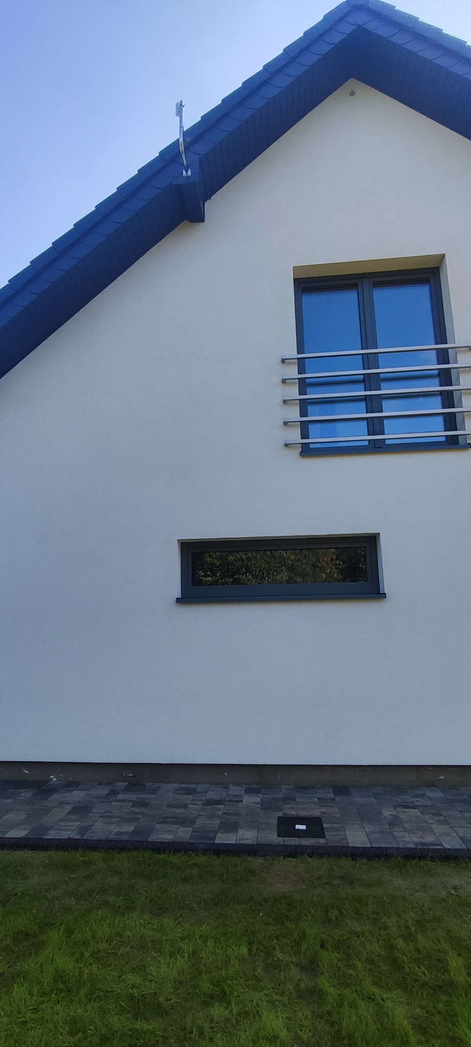 Biała elewacja domu z dwoma oknami i ciemną stolarką, widok z dołu na fragment budynku z kostką brukową i trawnikiem. Czysta, odświeżona powierzchnia.