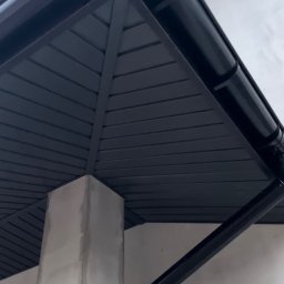 Strong-Bud - Ciemna podbitka dachowa z widocznym systemem rynnowym i fragmentem betonowej kolumny, ujęcie z dołu, podkreślające geometrię konstrukcji.