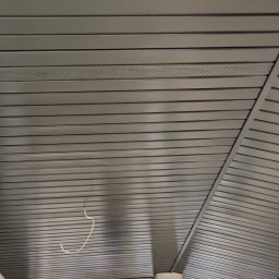 Strong-Bud - Szara podbitka dachowa z widocznymi perforacjami wentylacyjnymi, zamontowana na zewnątrz budynku. Widoczny fragment słupa i zarys drzew w tle.