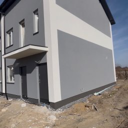 Strong-Bud - Dwukondygnacyjny dom z szarą elewacją i białym pasem, widoczny z boku, z ciemną podbitką dachową i dwoma wejściami. Teren wokół domu niezagospodarowany.