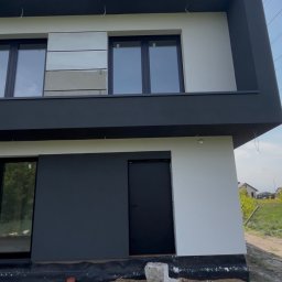 Strong-Bud - Dwukondygnacyjny dom z płaskim dachem, pomalowany na biało i grafitowo. Widoczne okna z czarnymi ramami i fragment terenu przed budynkiem.