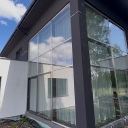 Strong-Bud - Nowoczesny dom z dużymi oknami i ciemną elewacją, widok z zewnątrz. Detal wykończenia przy gruncie. Błękitne niebo z chmurami odbija się w szybach.