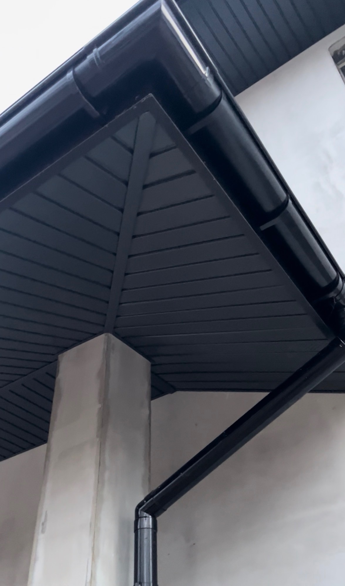 Ciemna podbitka dachowa z widocznym systemem rynnowym i fragmentem betonowej kolumny, ujęcie z dołu, podkreślające geometrię konstrukcji.