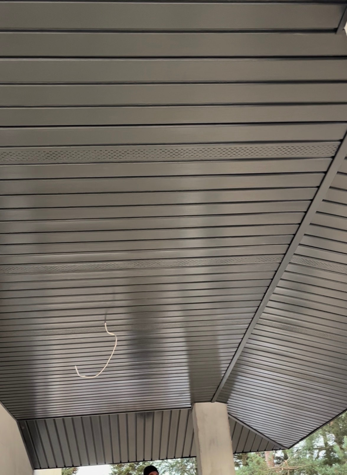 Szara podbitka dachowa z widocznymi perforacjami wentylacyjnymi, zamontowana na zewnątrz budynku. Widoczny fragment słupa i zarys drzew w tle.