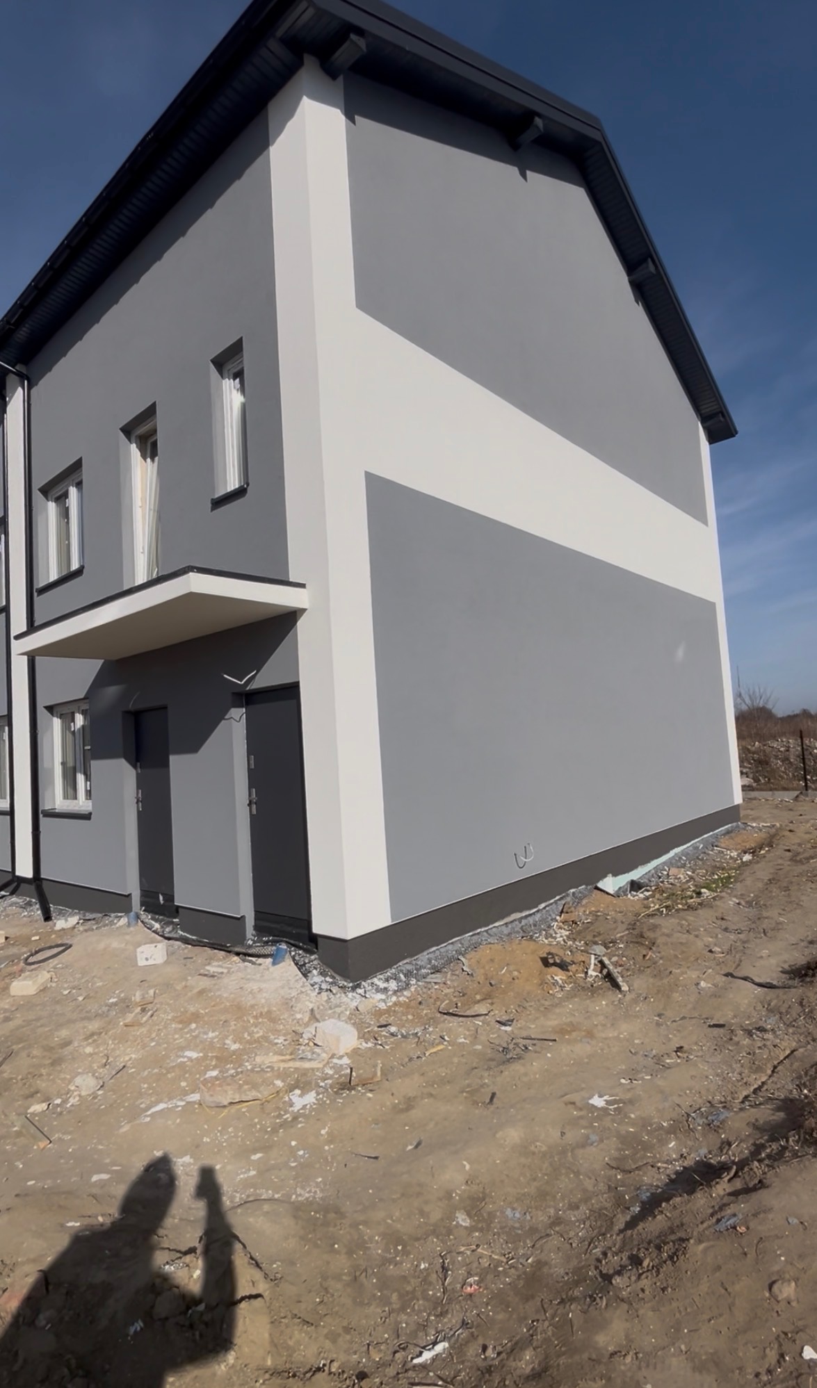 Dwukondygnacyjny dom z szarą elewacją i białym pasem, widoczny z boku, z ciemną podbitką dachową i dwoma wejściami. Teren wokół domu niezagospodarowany.