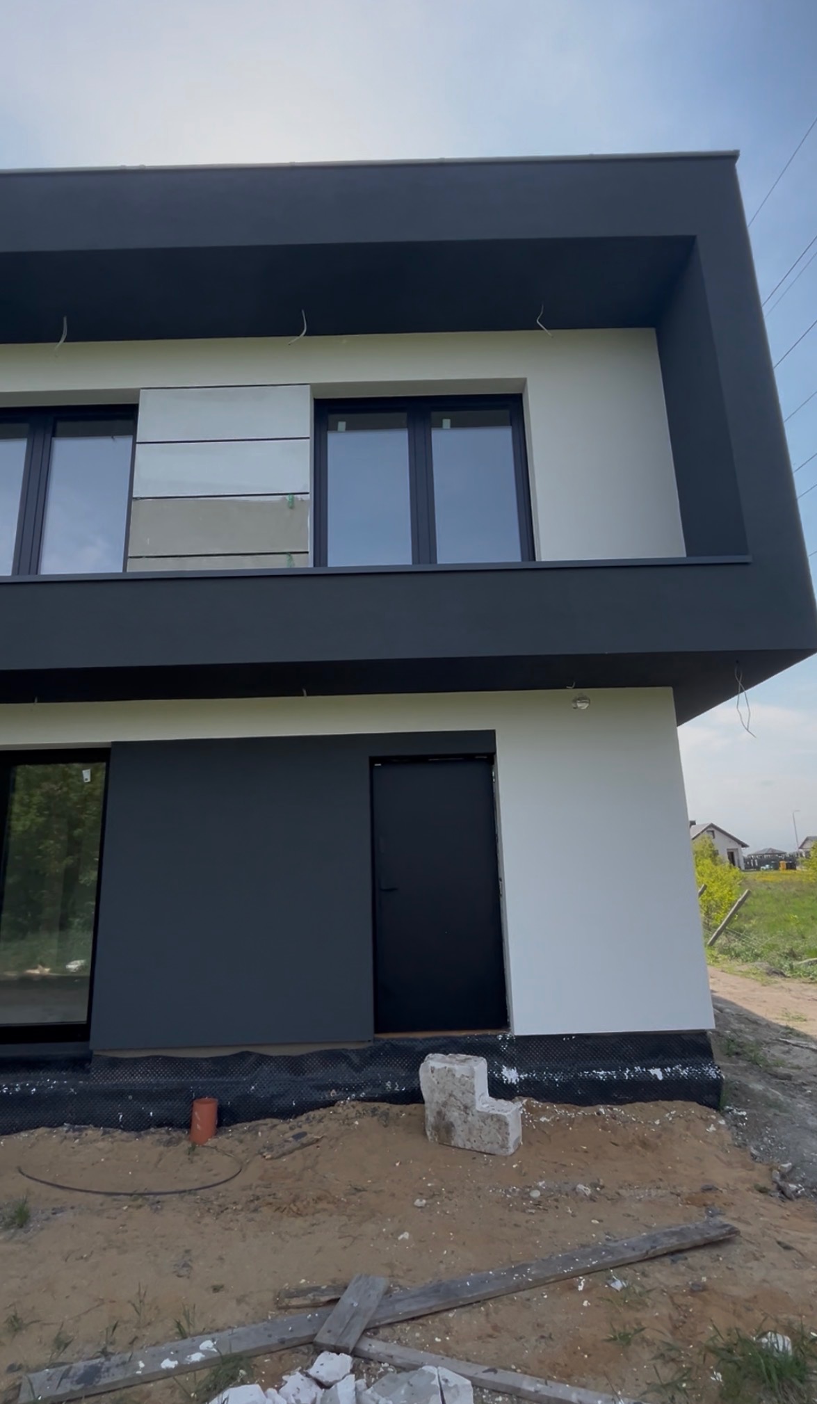 Dwukondygnacyjny dom z płaskim dachem, pomalowany na biało i grafitowo. Widoczne okna z czarnymi ramami i fragment terenu przed budynkiem.
