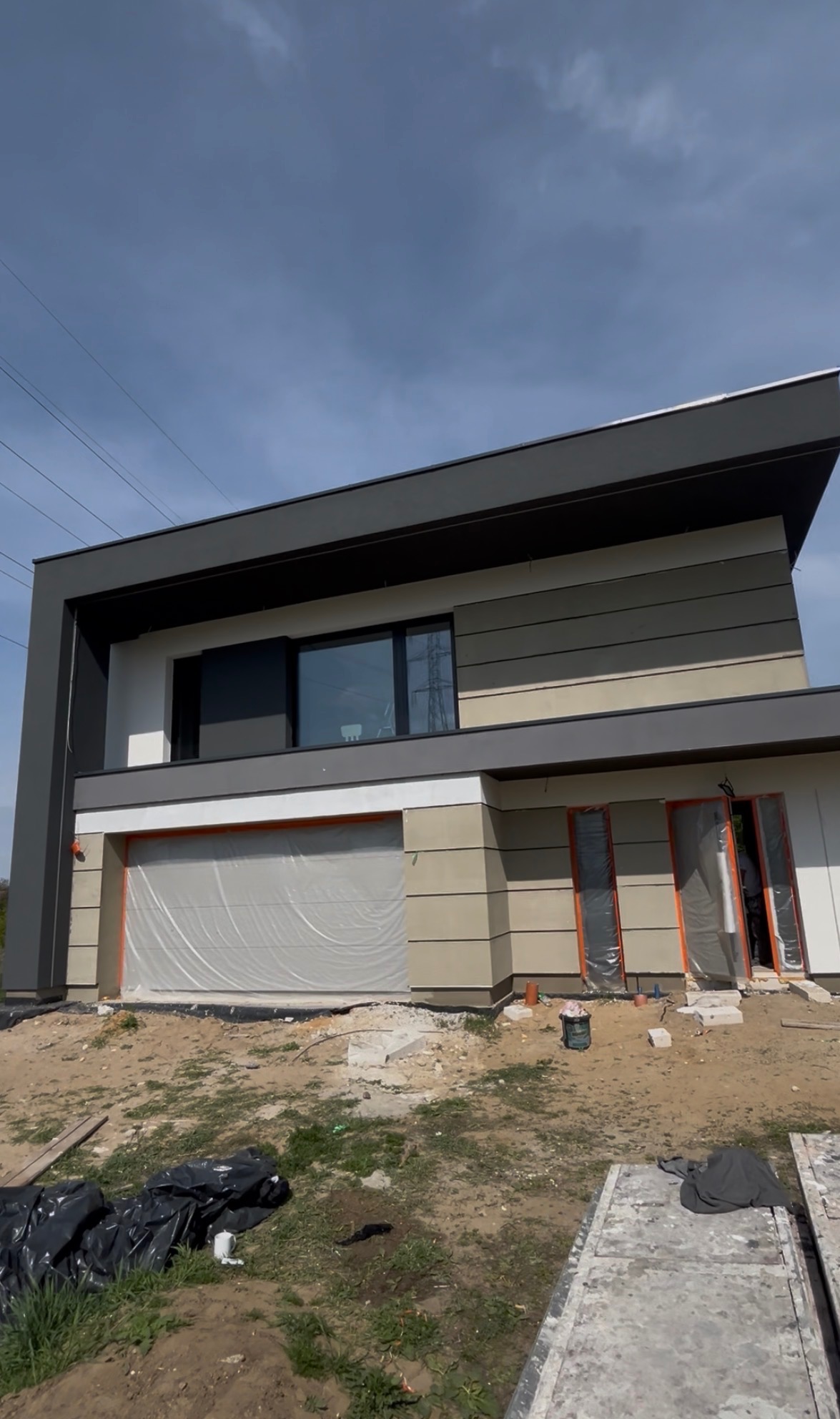 Nowoczesny dom z elewacją w odcieniach szarości i beżu, okna i brama garażowa zabezpieczone folią, teren wokół w trakcie prac wykończeniowych.