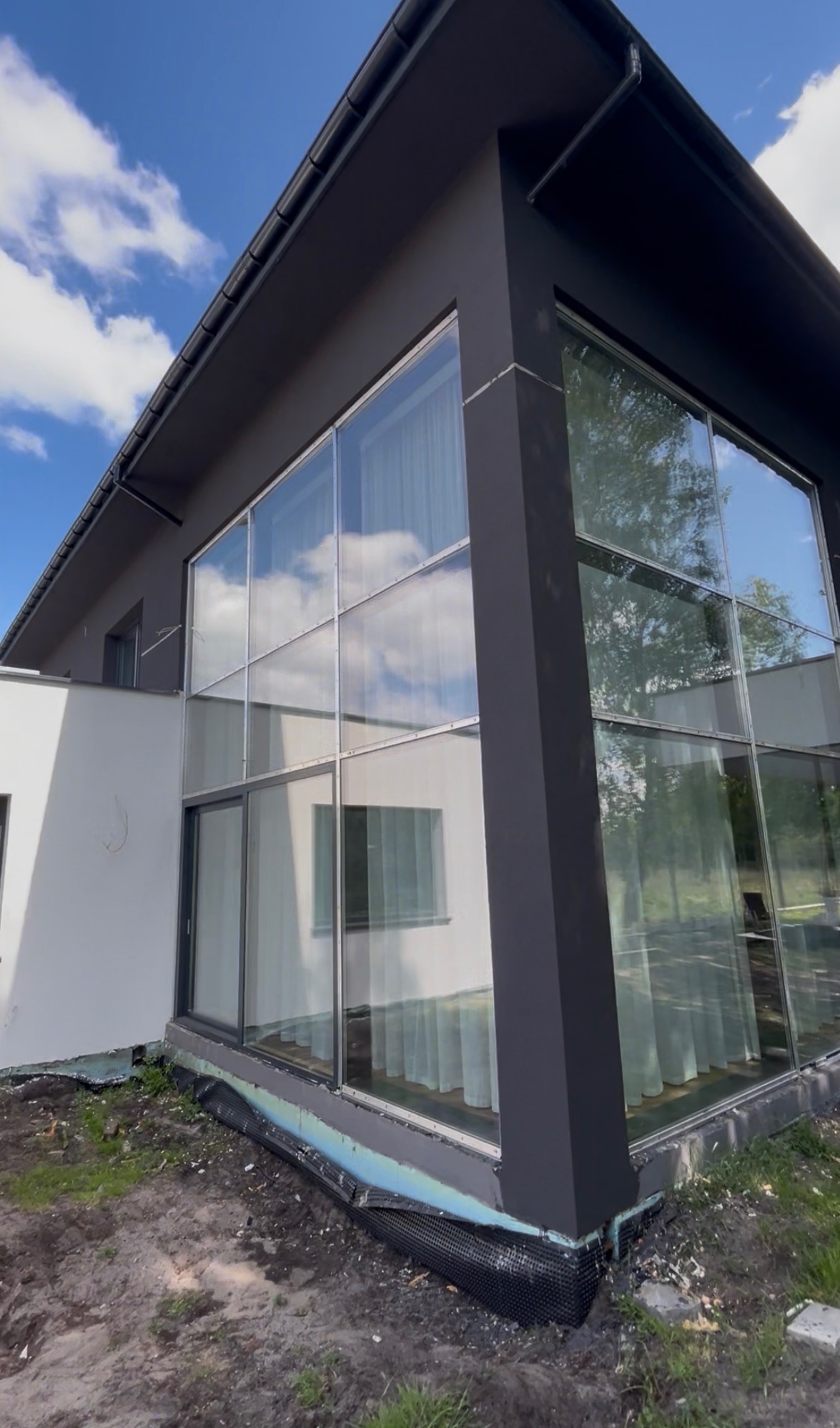 Nowoczesny dom z dużymi oknami i ciemną elewacją, widok z zewnątrz. Detal wykończenia przy gruncie. Błękitne niebo z chmurami odbija się w szybach.