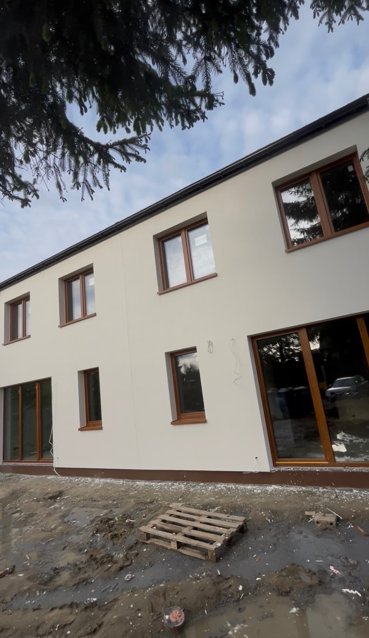 Jasna elewacja dwukondygnacyjnego domu z brązowymi ramami okiennymi. Na pierwszym planie błotniste podłoże i drewniana paleta. Widok z dołu, fragment drzewa w górnej części kadru.
