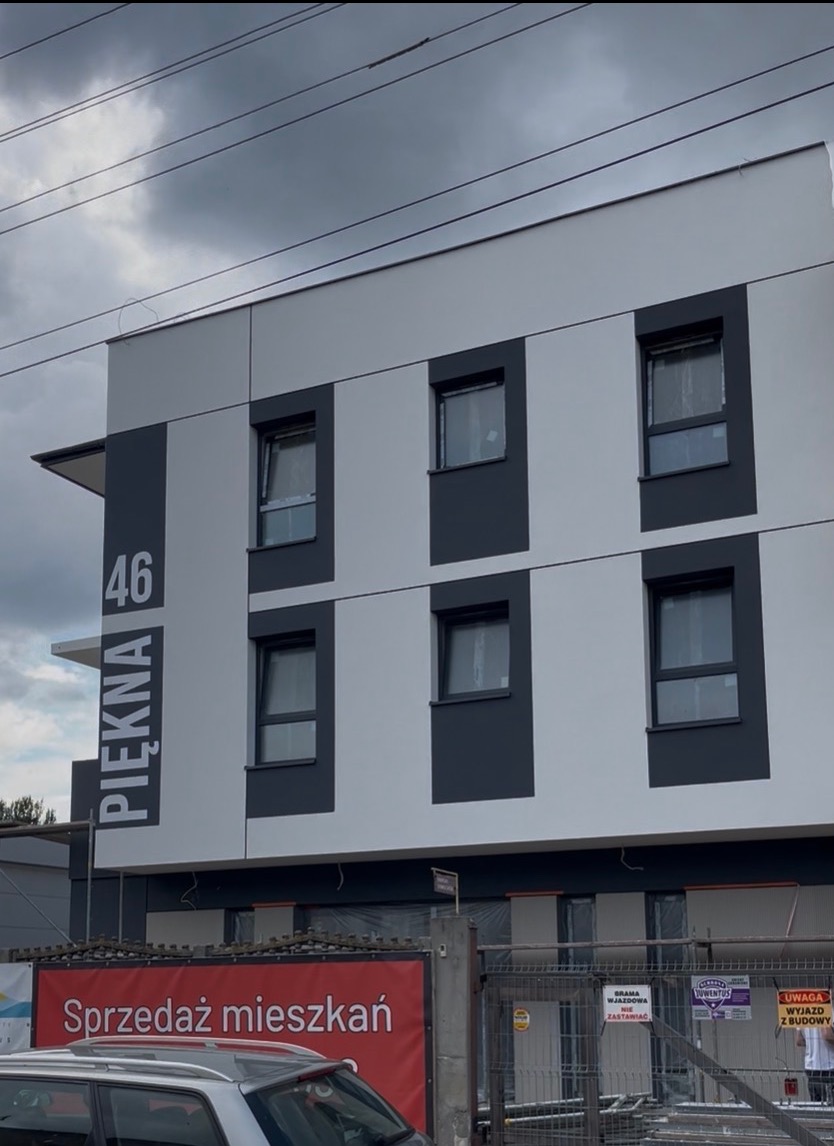 Nowoczesna elewacja budynku z szarymi akcentami i dużym napisem 'Piękna 46'. Widoczny fragment baneru 'Sprzedaż mieszkań' i zaparkowany samochód.