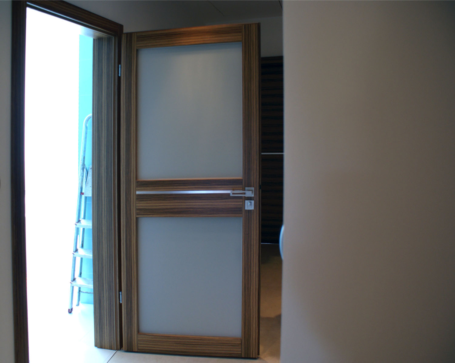 Otwarta, nowoczesna, wewnętrzna stolarka drzwiowa z drewnianą ramą i matowym szkłem, widoczna drabina w tle.