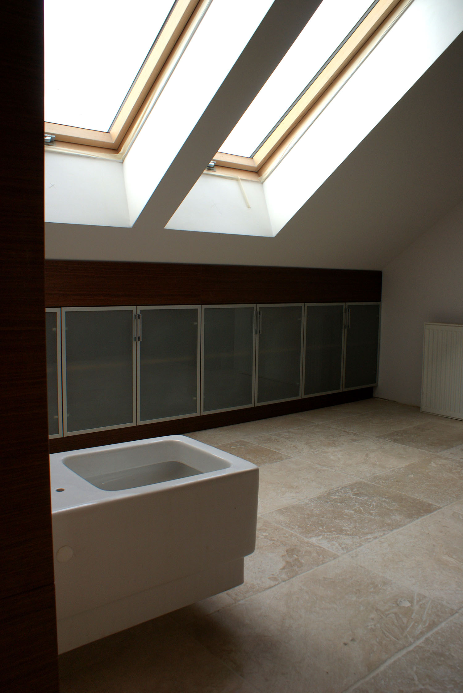 Poddasze ze skośnym sufitem doświetlone trzema oknami dachowymi. W tle zabudowa szafek z ciemnego drewna i szarymi, matowymi frontami. Na pierwszym planie biała wanna z hydromasażem.