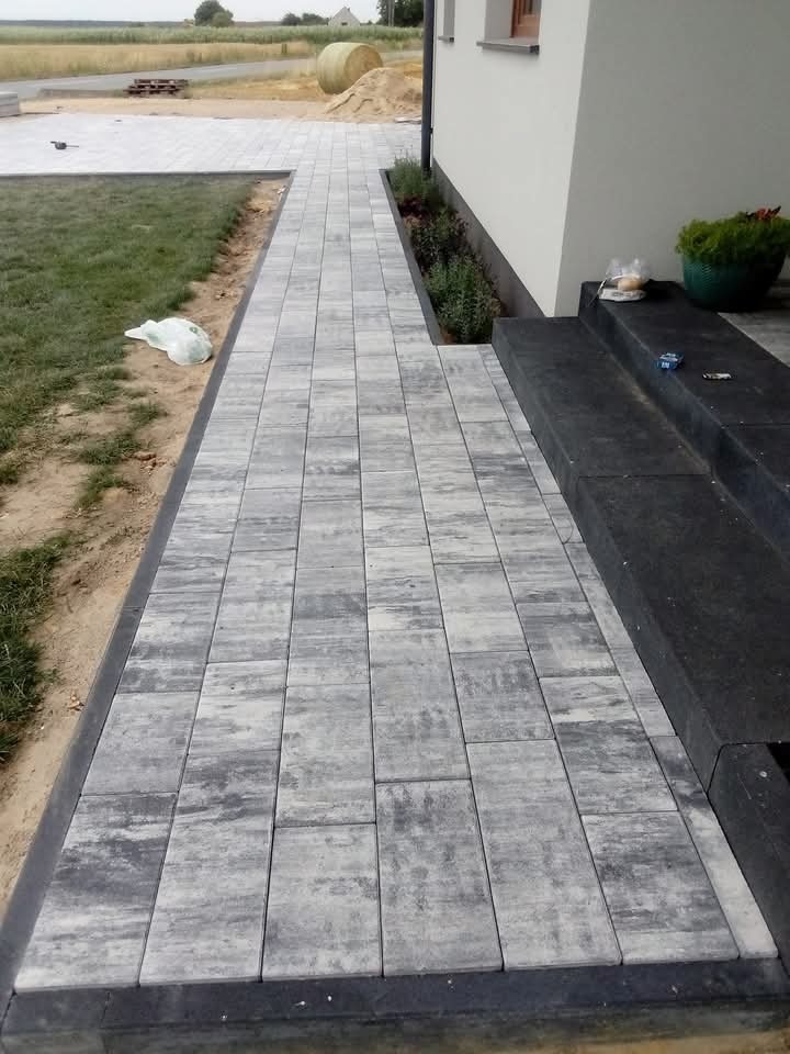 Ścieżka z szarej kostki brukowej prowadząca do domu, z widocznym trawnikiem i elementami krajobrazu. Kostka ułożona równo, z czarnym obrzeżem. W tle balot słomy.