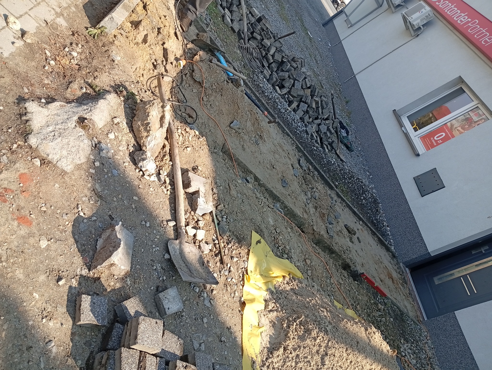 Przygotowanie podłoża pod kostkę brukową: wykop, piasek, narzędzia (łopata, grabie), obrzeża i kostka. Widoczny budynek z oknem i szyldem Santander Partner.