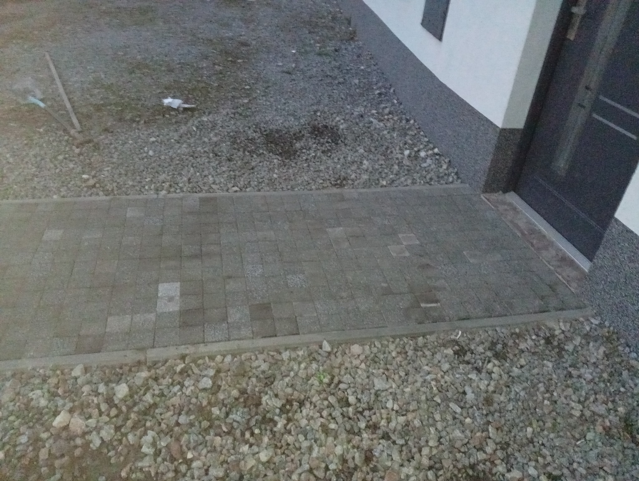 Szara kostka brukowa prowadząca do drzwi wejściowych domu, otoczona kamieniami. Widoczne fragmenty elewacji i drzwi. Ujęcie z góry, lekko z boku.
