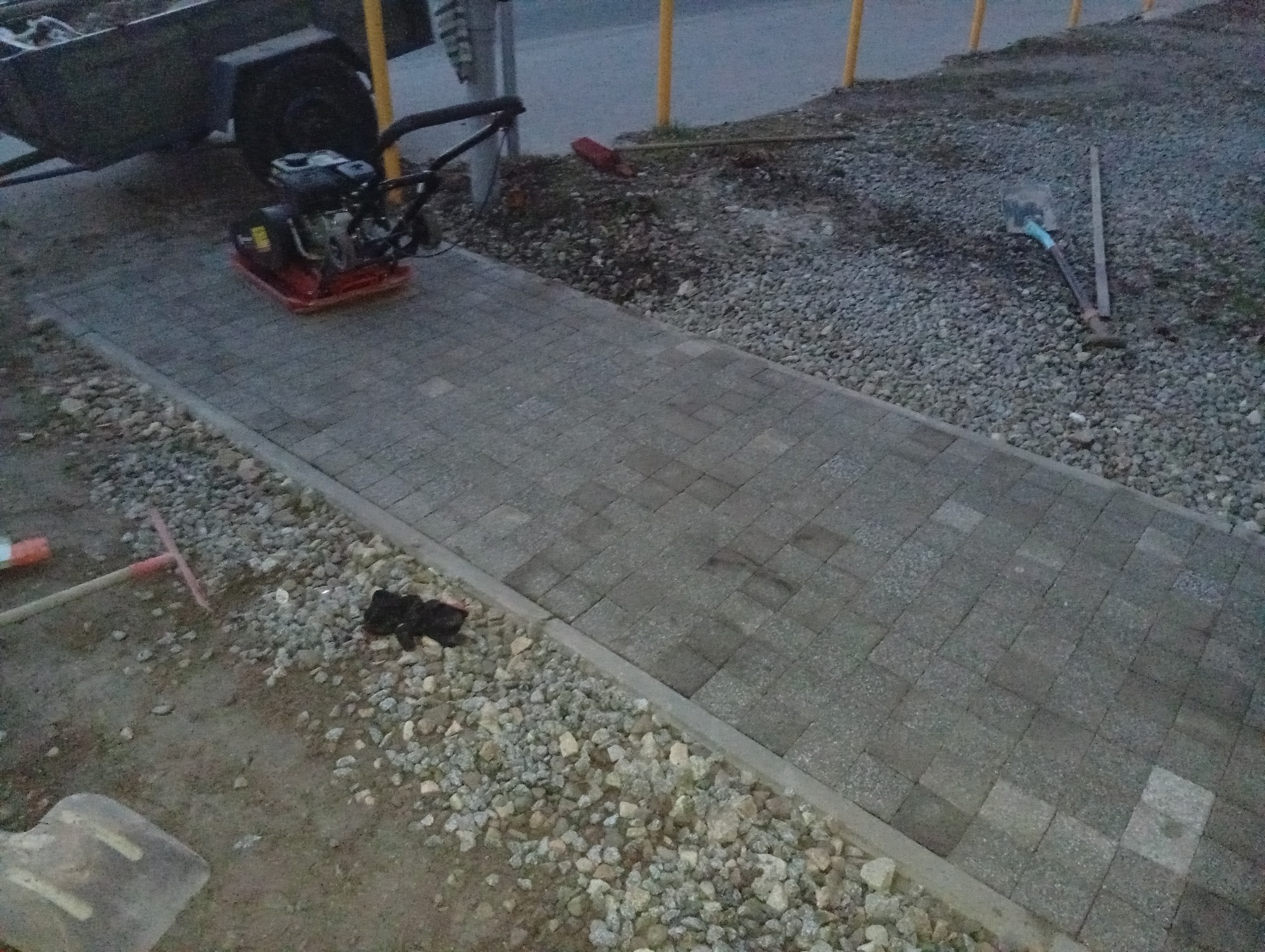 Świeżo ułożona ścieżka z kostki brukowej w odcieniach szarości, z zagęszczarką w tle i narzędziami obok, otoczona żwirem.