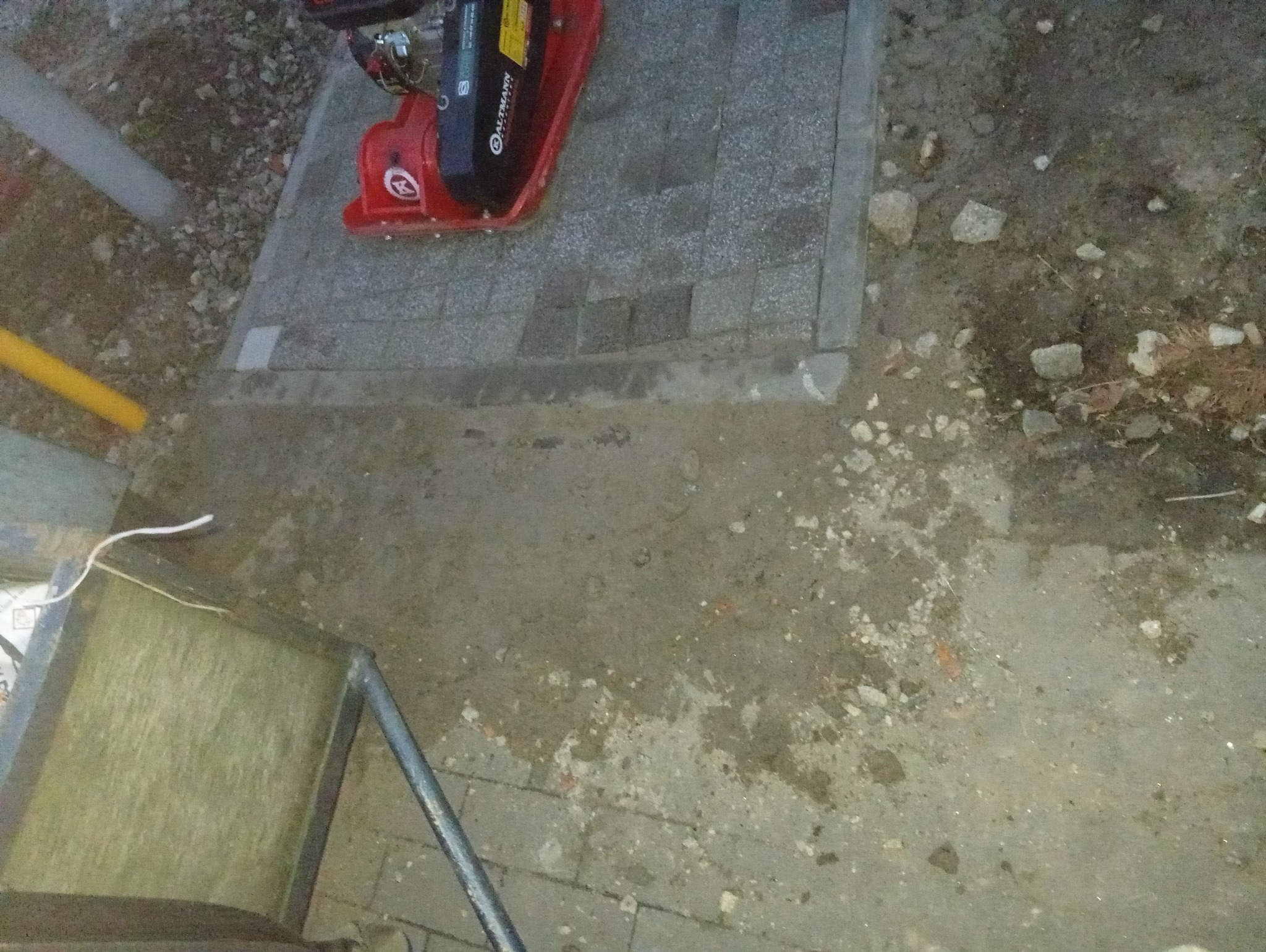 Częściowo ułożona kostka brukowa z zagęszczarką w rogu, widoczne przygotowane podłoże i elementy otoczenia. Nierówności terenu i niedokończony etap prac.