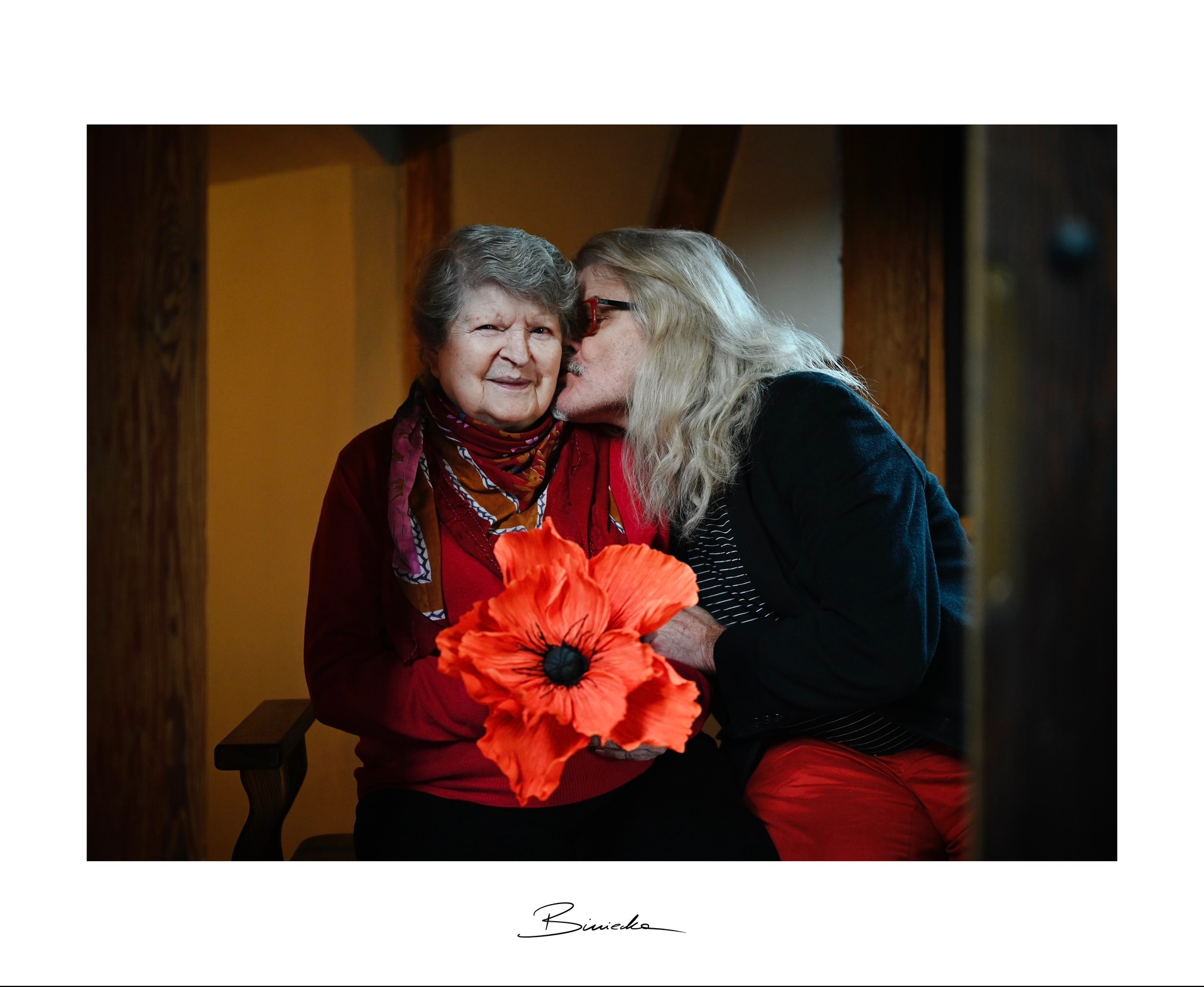 Portret uśmiechniętej starszej kobiety z dużym, czerwonym makiem, całowanej w policzek przez drugą kobietę w okularach i czarnej marynarce. Ciepłe, naturalne światło.