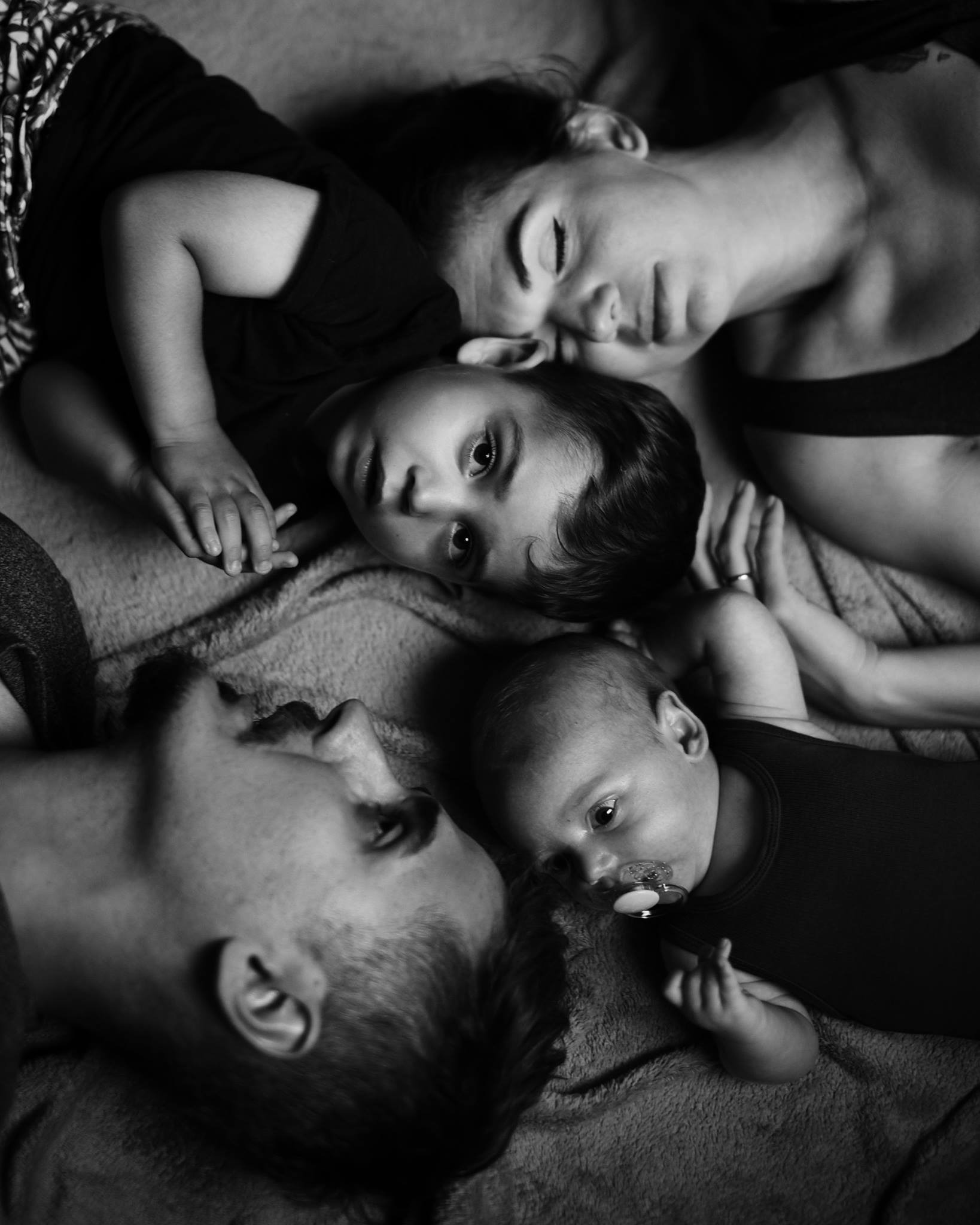 Czarno-białe, artystyczne ujęcie rodziny leżącej blisko siebie: ojciec z brodą, matka z zamkniętymi oczami, starsze dziecko patrzące w obiektyw i niemowlę z smoczkiem.