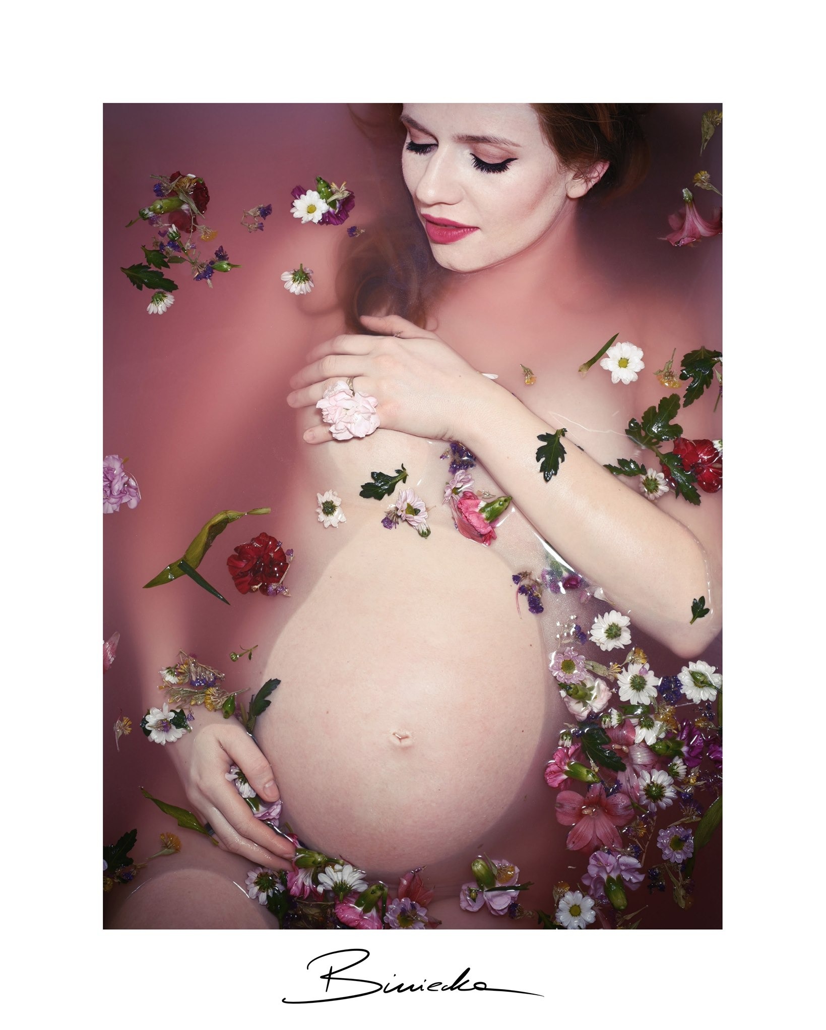 Kreatywne ujęcie kobiety w ciąży, zanurzonej w wodzie z kwiatami, z delikatnym oświetleniem i subtelnym makijażem, podkreślające piękno oczekiwania na dziecko.