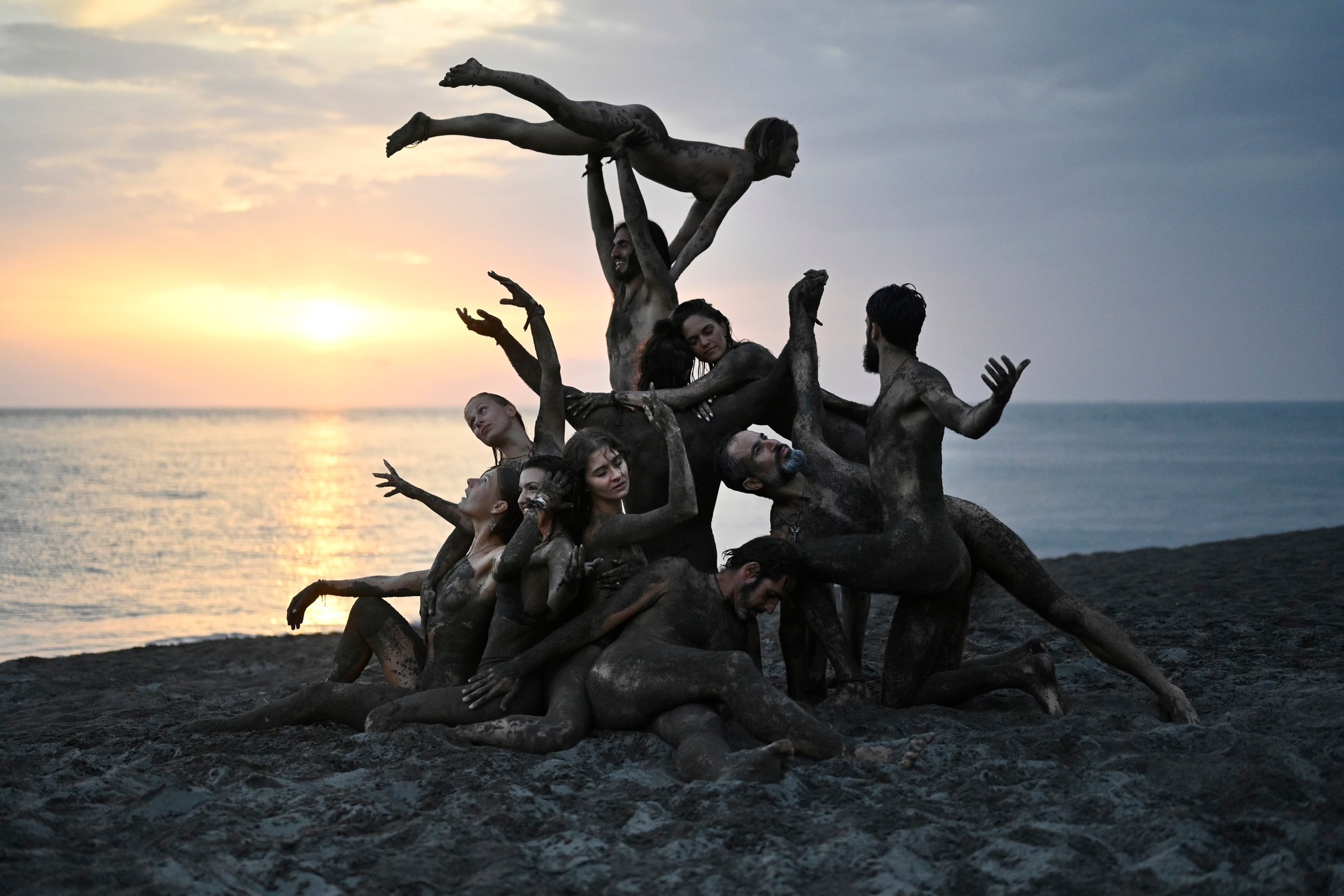 Dynamiczna kompozycja grupy ludzi pokrytych gliną na plaży o zachodzie słońca. Artystyczne ujęcie z elementami akrobatyki i ekspresji ciała na tle morza.