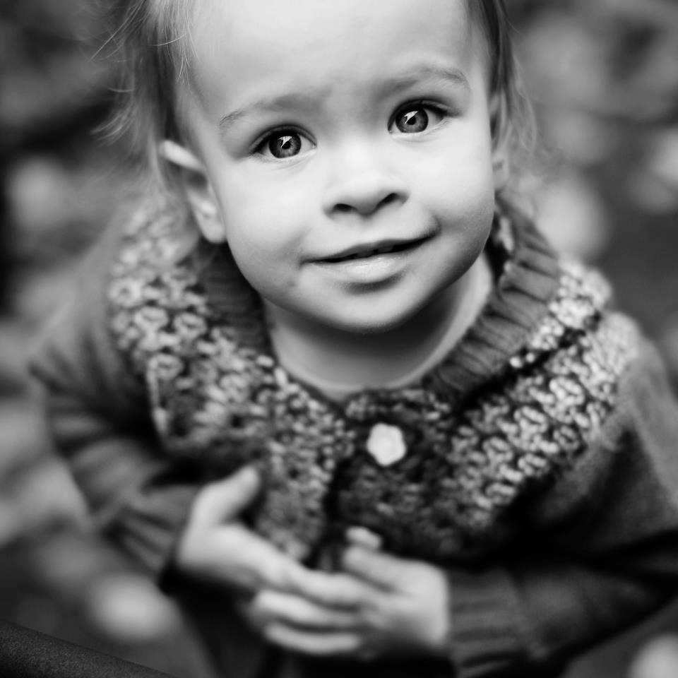 Czarno-białe, portretowe ujęcie od dołu uśmiechniętej dziewczynki w swetrze z dużym splotem, z delikatnym rozmyciem tła.