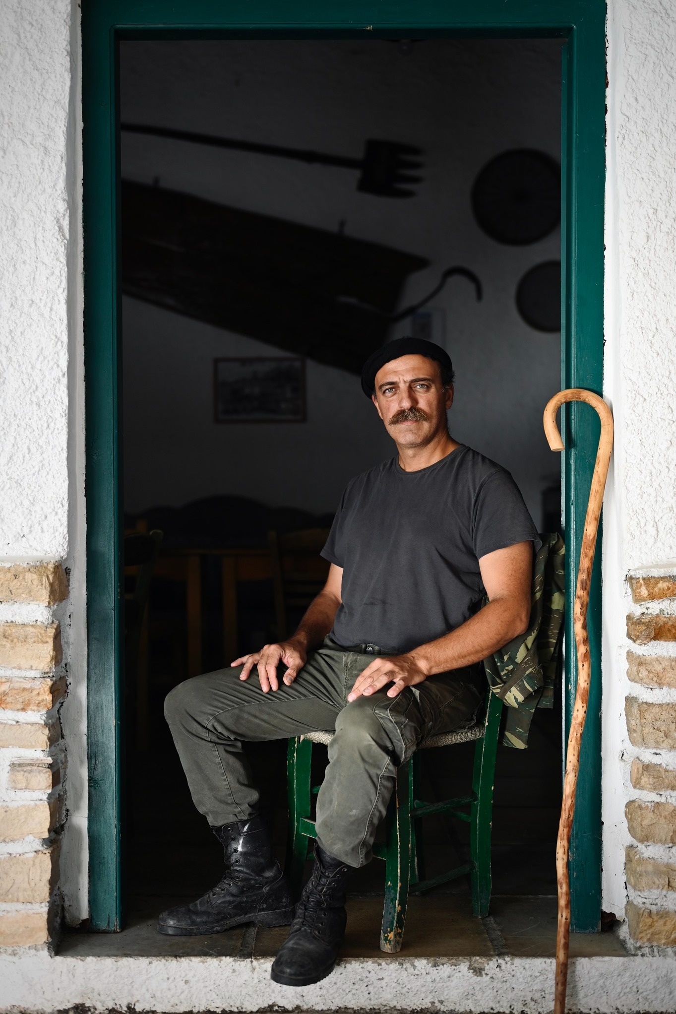 Mężczyzna z wąsami w berecie siedzi na krześle w drzwiach, opierając się o laskę. W tle ciemne wnętrze z narzędziami rolniczymi. Ujęcie portretowe.