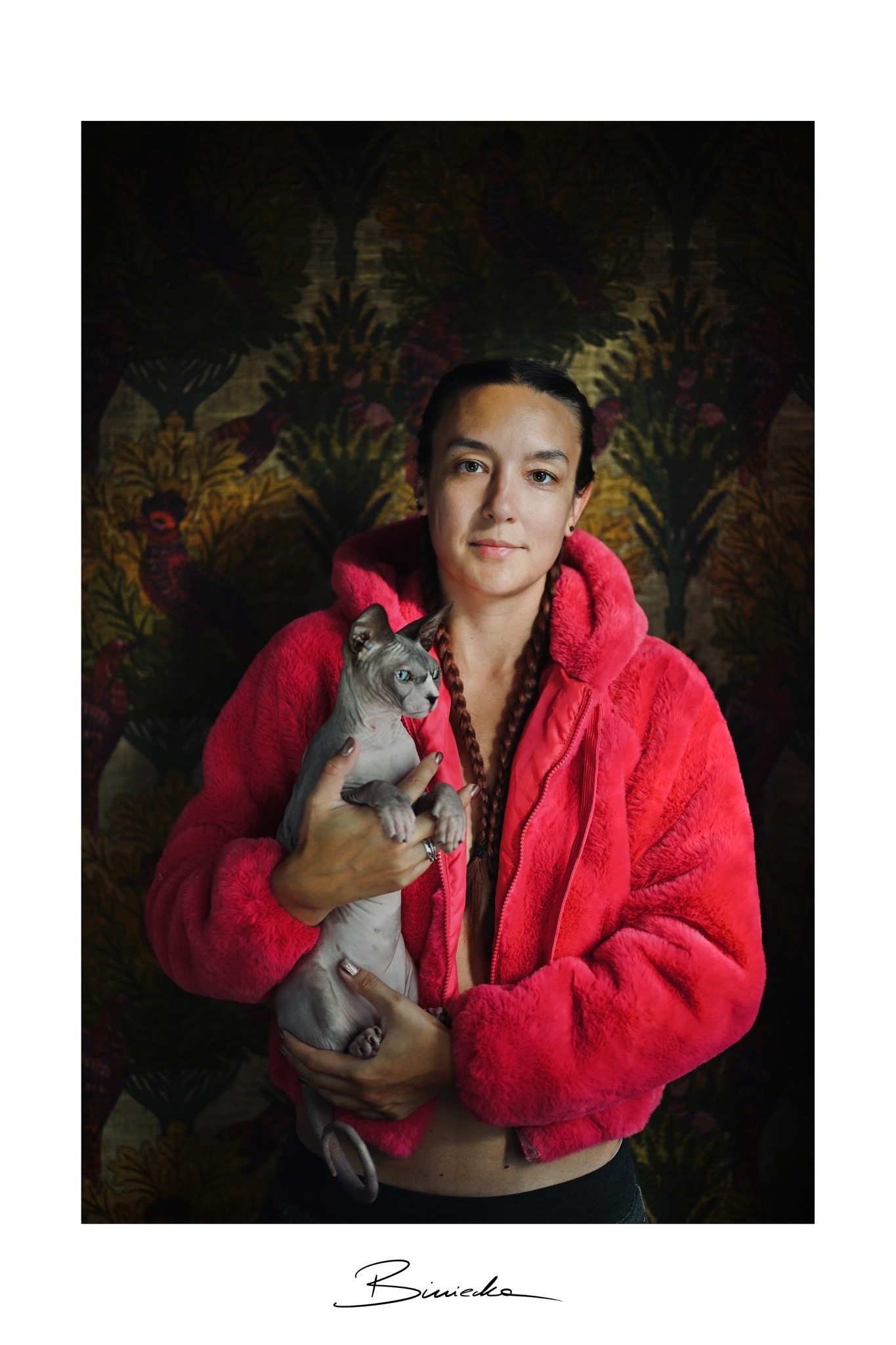 Portret kobiety w różowej kurtce z kapturem, trzymającej kota rasy sfinks na tle wzorzystej tapety. Uwagę przykuwają warkocze i wyraz twarzy modelki.