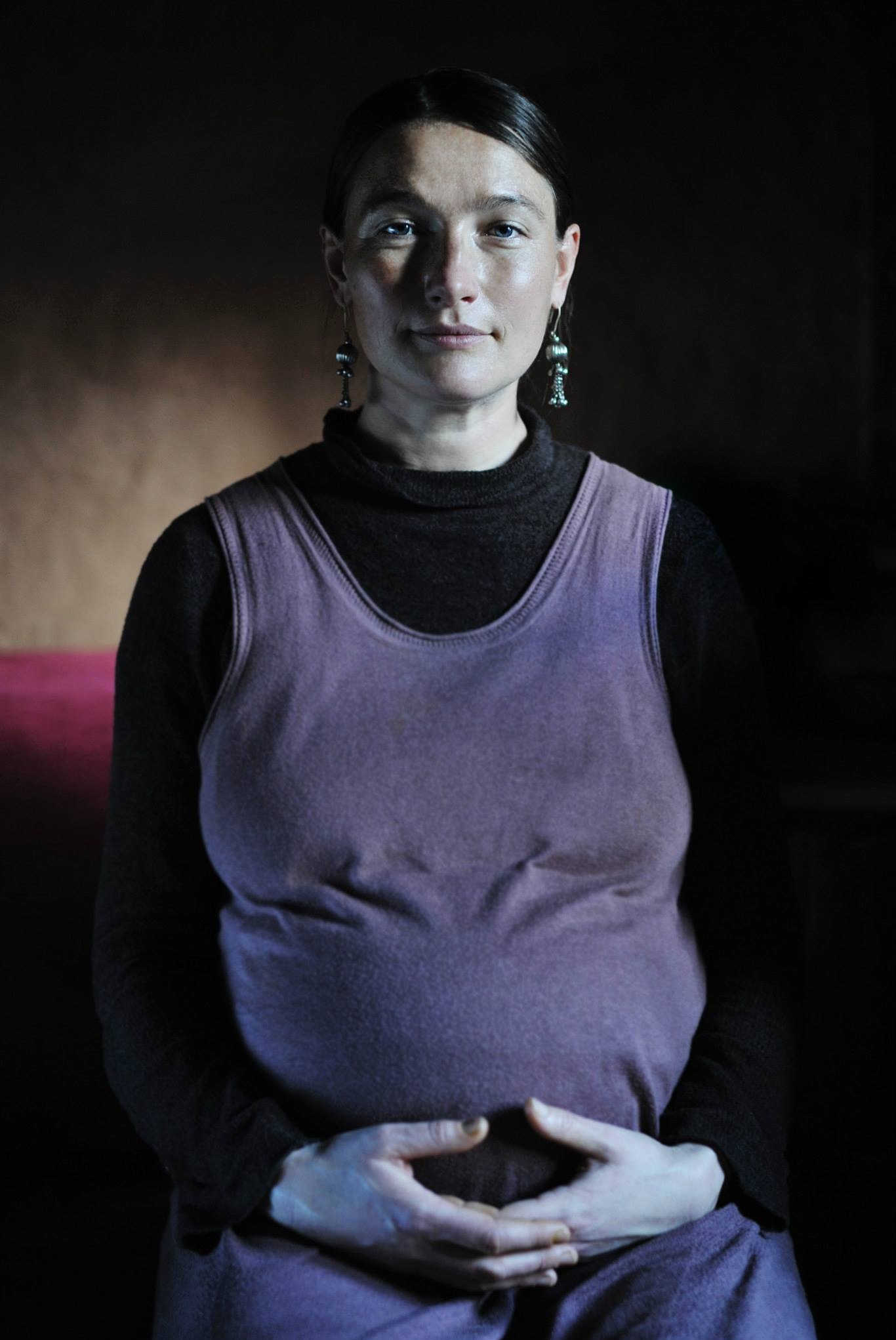 Portret kobiety w ciąży, ujęcie centralne, ciemne tło, ręce splecione na brzuchu, kolczyki w uszach, fioletowa sukienka na czarnym golfie. Subtelne światło podkreśla rysy twarzy.