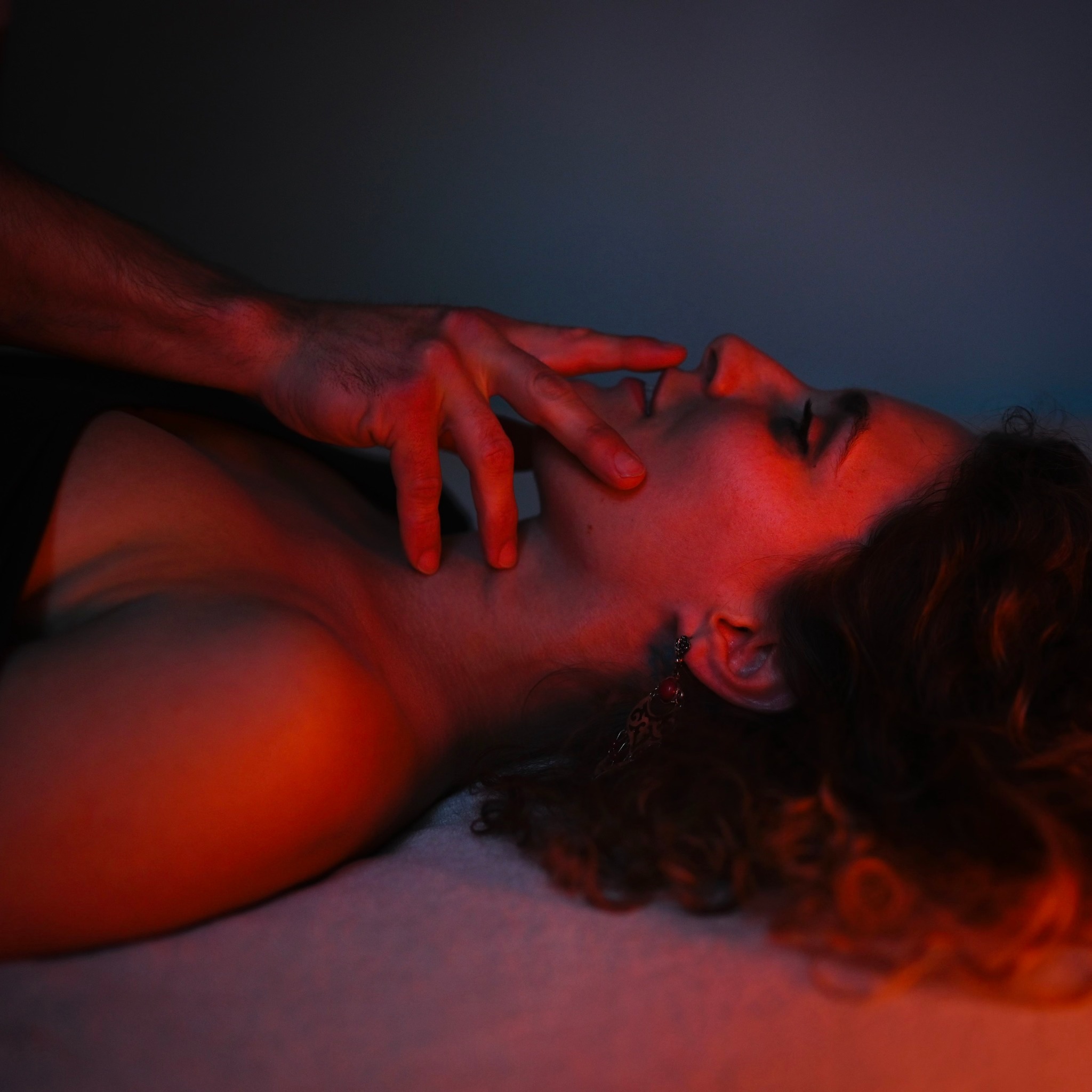 Kobieta leży na plecach, oświetlona czerwonym światłem, z dłonią mężczyzny dotykającą jej twarzy. Widoczne kolczyki i fragment czarnego ubrania.