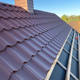 Usługi dekarsko-ciesielskie Kacper Szadkowski - Montaż brązowej blachodachówki trapezowej z kominem na dachu domu jednorodzinnego. Widoczna konstrukcja drewniana i membrana dachowa. Tło stanowi błękitne niebo i pola.