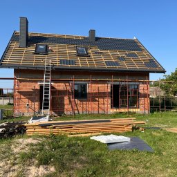 Usługi dekarsko-ciesielskie Kacper Szadkowski - Dom w trakcie budowy z częściowo pokrytym dachem czarną dachówką, widoczne rusztowania, drewno i okna dachowe. Ujęcie w słoneczny dzień.