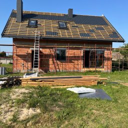 Usługi dekarsko-ciesielskie Kacper Szadkowski - Budowa domu jednorodzinnego w trakcie krycia dachu blachodachówką w kolorze grafitowym. Widoczne rusztowania, drabina i materiały budowlane. Słoneczny dzień.