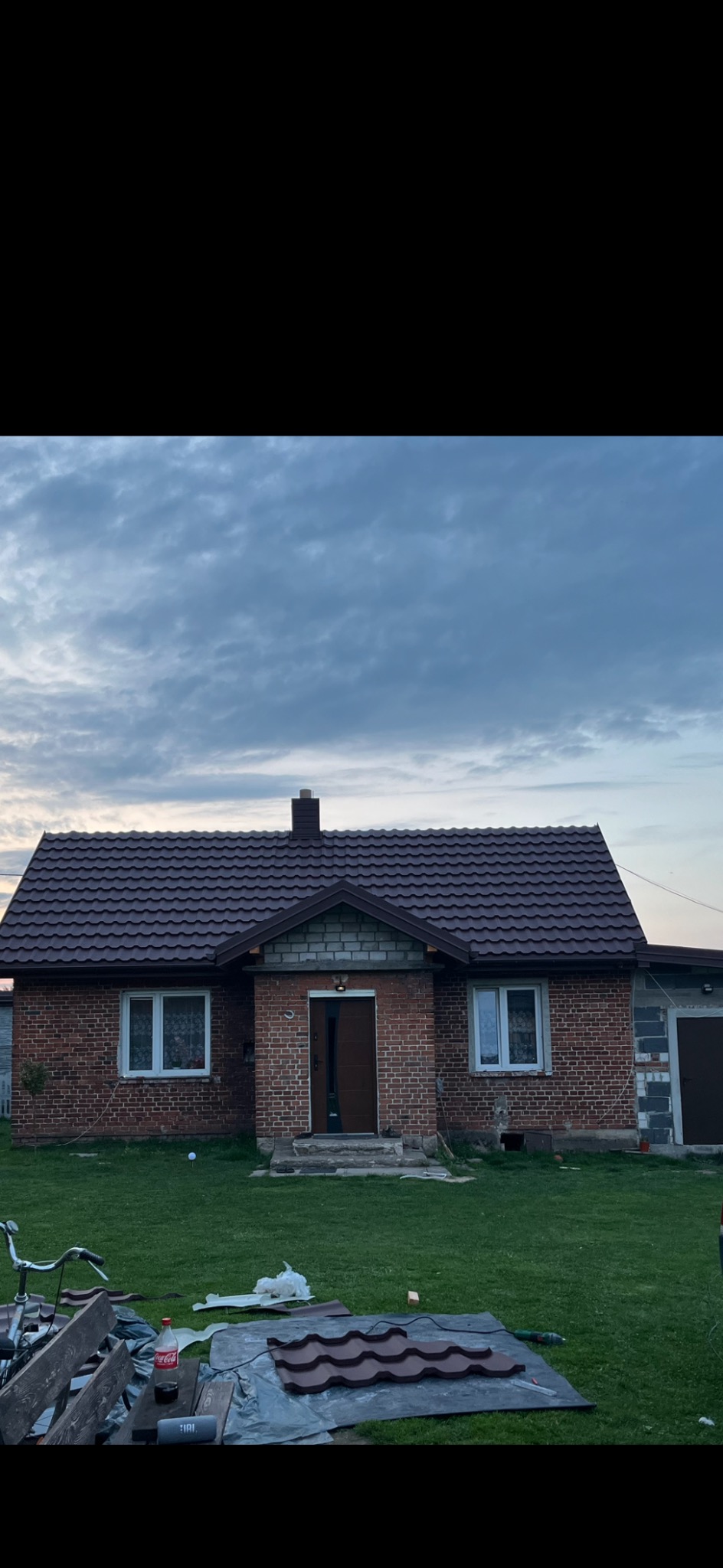 Dom z nowym, brązowym dachem z blachodachówki w Sieradzu. Na pierwszym planie arkusze blachy przygotowane do montażu. Widok na budynek z ogrodu.