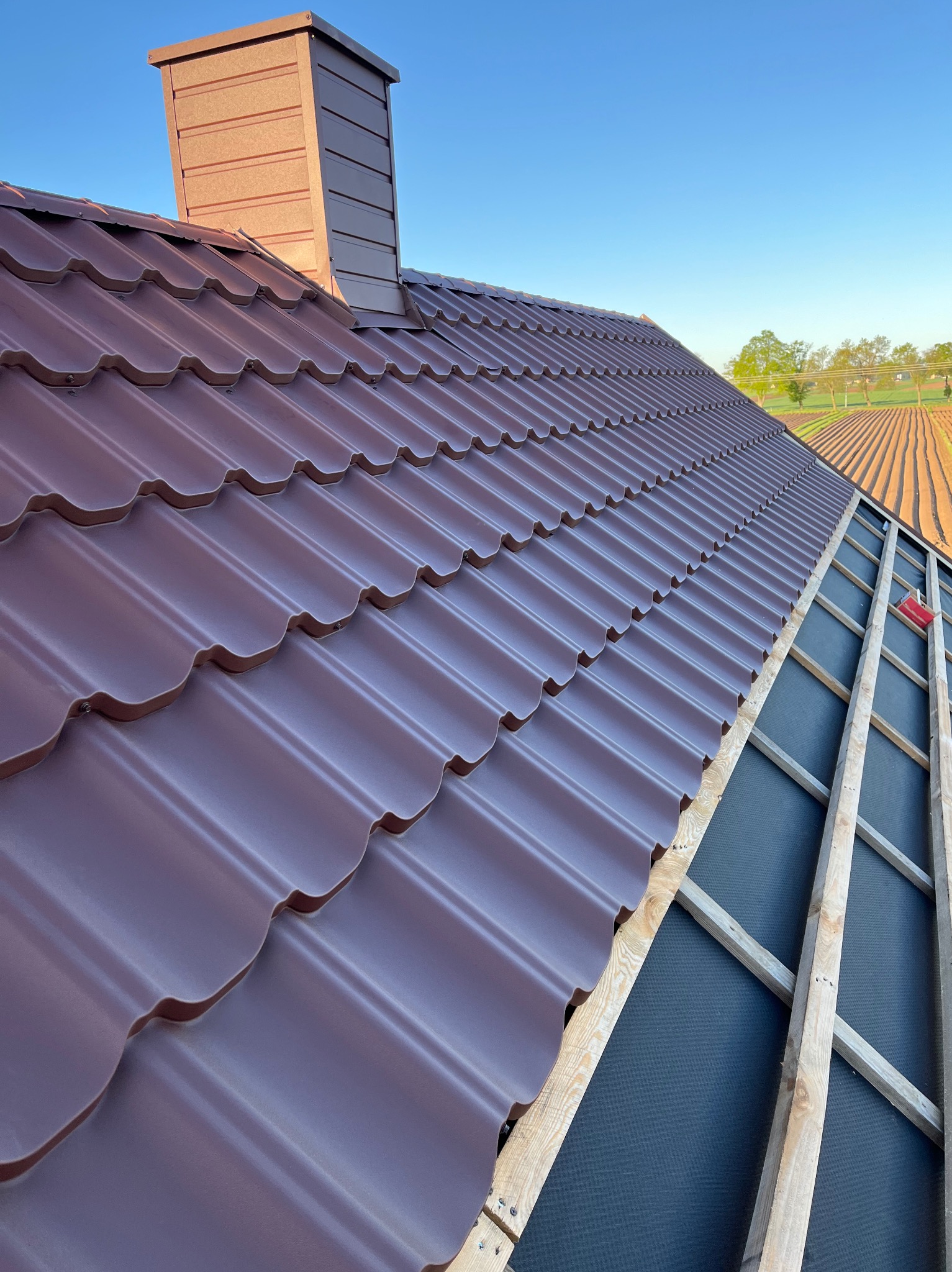 Montaż brązowej blachodachówki trapezowej z kominem na dachu domu jednorodzinnego. Widoczna konstrukcja drewniana i membrana dachowa. Tło stanowi błękitne niebo i pola.