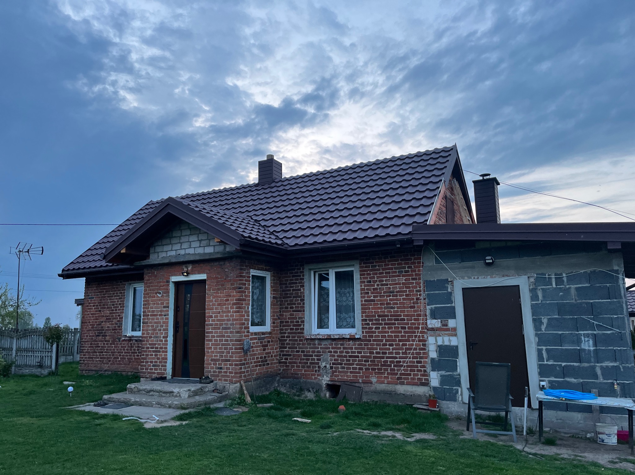 Dom z brązowym dachem z blachy trapezowej, ceglaną elewacją i dobudówką z bloczków. Widoczne kominy i pochmurne niebo. Ujęcie z poziomu gruntu.